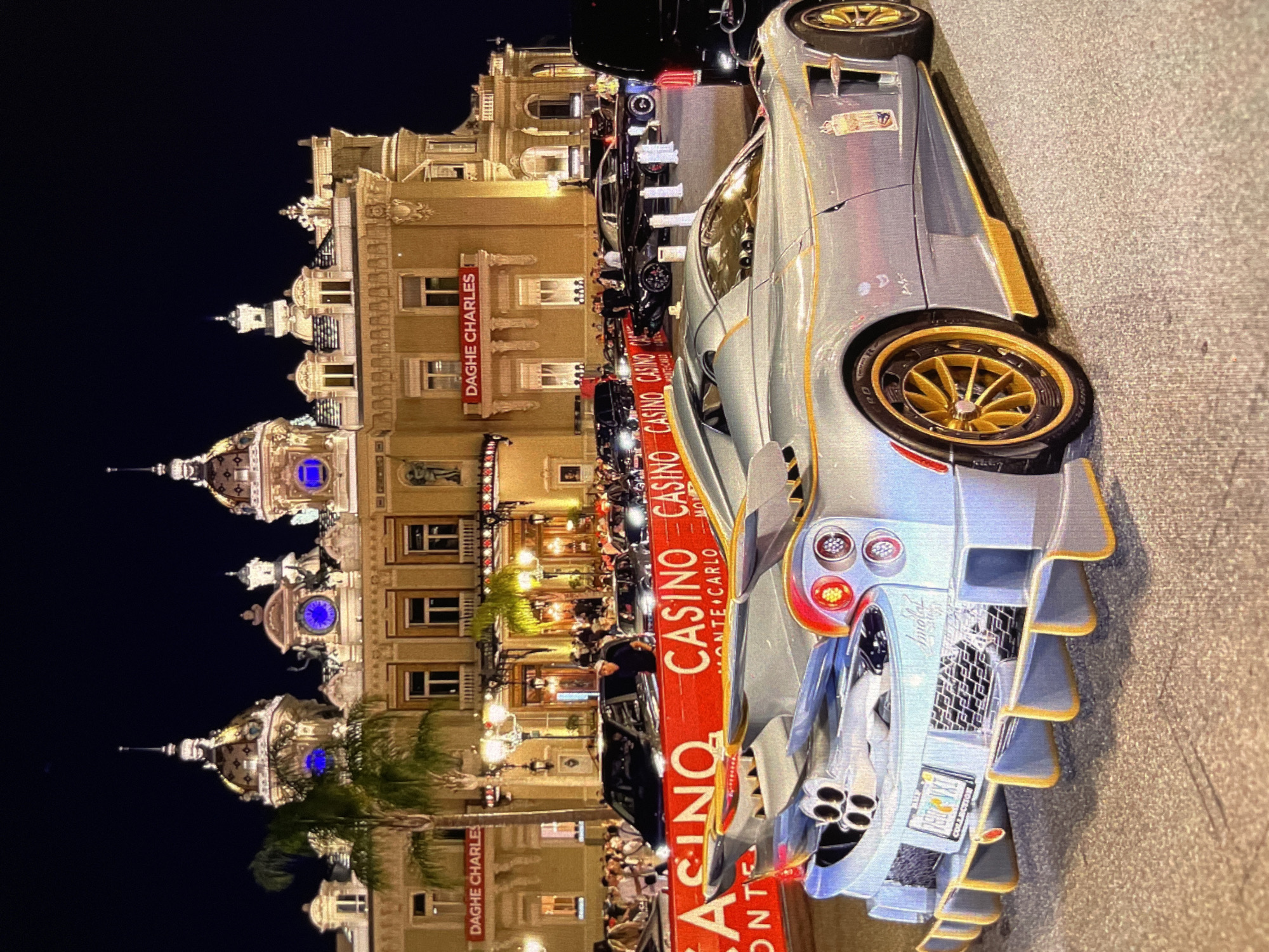 At nighttime, a light blue sports car with gold accents is parked on a paved street in front of a large, ornate building. The building, which appears to be a casino, is illuminated with lights and features classical architecture with domes and spires. Red banners with white lettering that reads "CASINO MONTE CARLO" are displayed in front of the building. Several other cars and a crowd of people are visible in the background. The sports car has a distinctive rear spoiler and quad exhaust pipes.