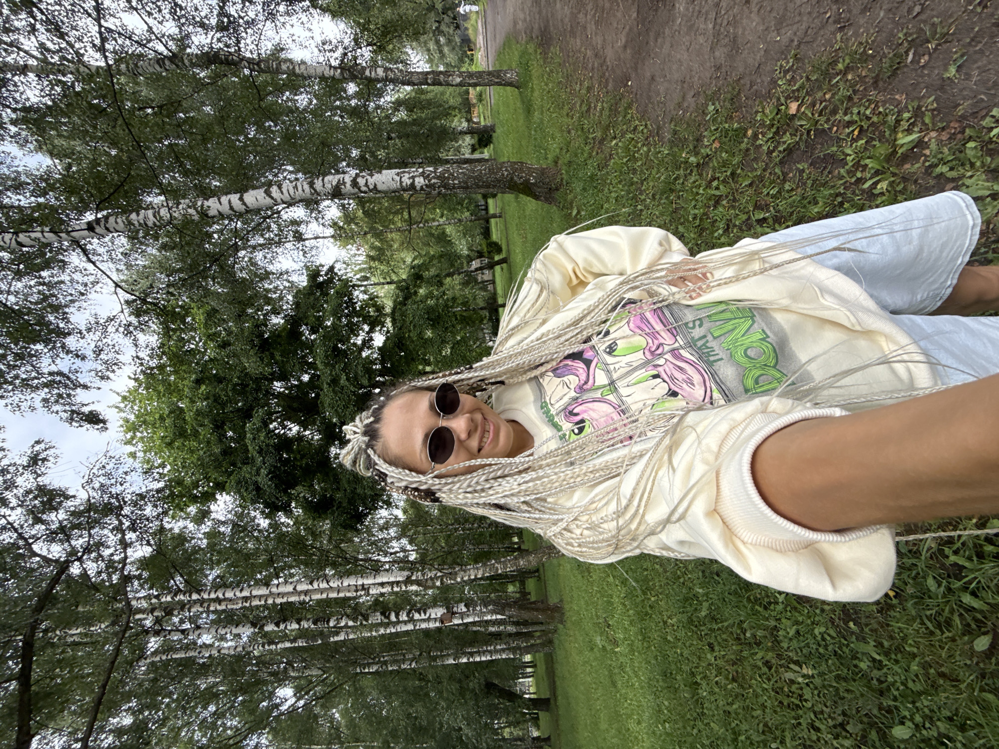 A young woman with blonde braided hair and sunglasses is taking a selfie in a park in Rusanovo, Russia. She is wearing a light yellow sweatshirt with a graphic print and light blue shorts. The park has birch trees, lush green grass, and a dirt path. The weather appears overcast.