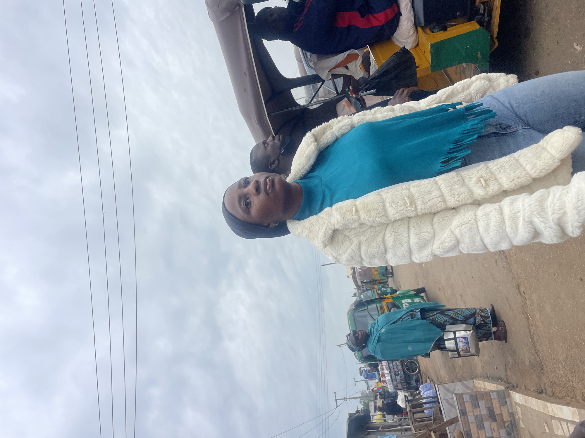 In Gura Top, Nigeria, a woman wearing a black hijab, a teal turtleneck with fringed detailing, and a white faux fur coat stands on a dirt road. She is looking to her left, her expression thoughtful. Behind her, a man in a dark top is also visible, wearing a black face mask. Further in the background, a yellow and green auto-rickshaw, also known as a "Keke," is parked. Another auto-rickshaw is in motion on the road. To the left of the frame, other individuals are visible, along with market stalls. An older woman in a teal headscarf and patterned dress is also present, carrying a silver tote bag. The sky above is overcast.