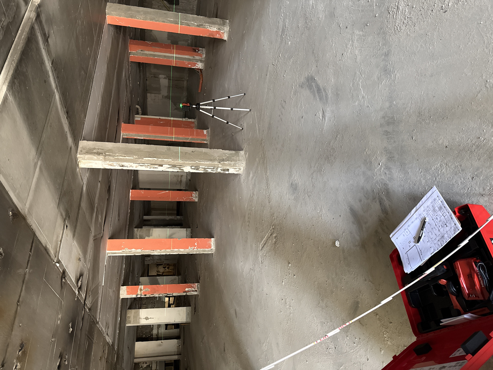 The image shows the interior of a construction site. It is an elevator shaft. The shaft walls are dark gray and are lined with metal. The shaft interior reveals several staggered rectangular supports protruding from the wall. These supports are painted orange-red and have concrete extensions on top.
A laser level tripod stands within the shaft, with green laser lines visible along the supports. A red toolbox lies on the ground, with architectural drawings visible on top of it. A white and red measuring tape extends from the toolbox.
The scene does not provide conclusive details or visual references that would allow for identification of the location as Varna, Bulgaria.