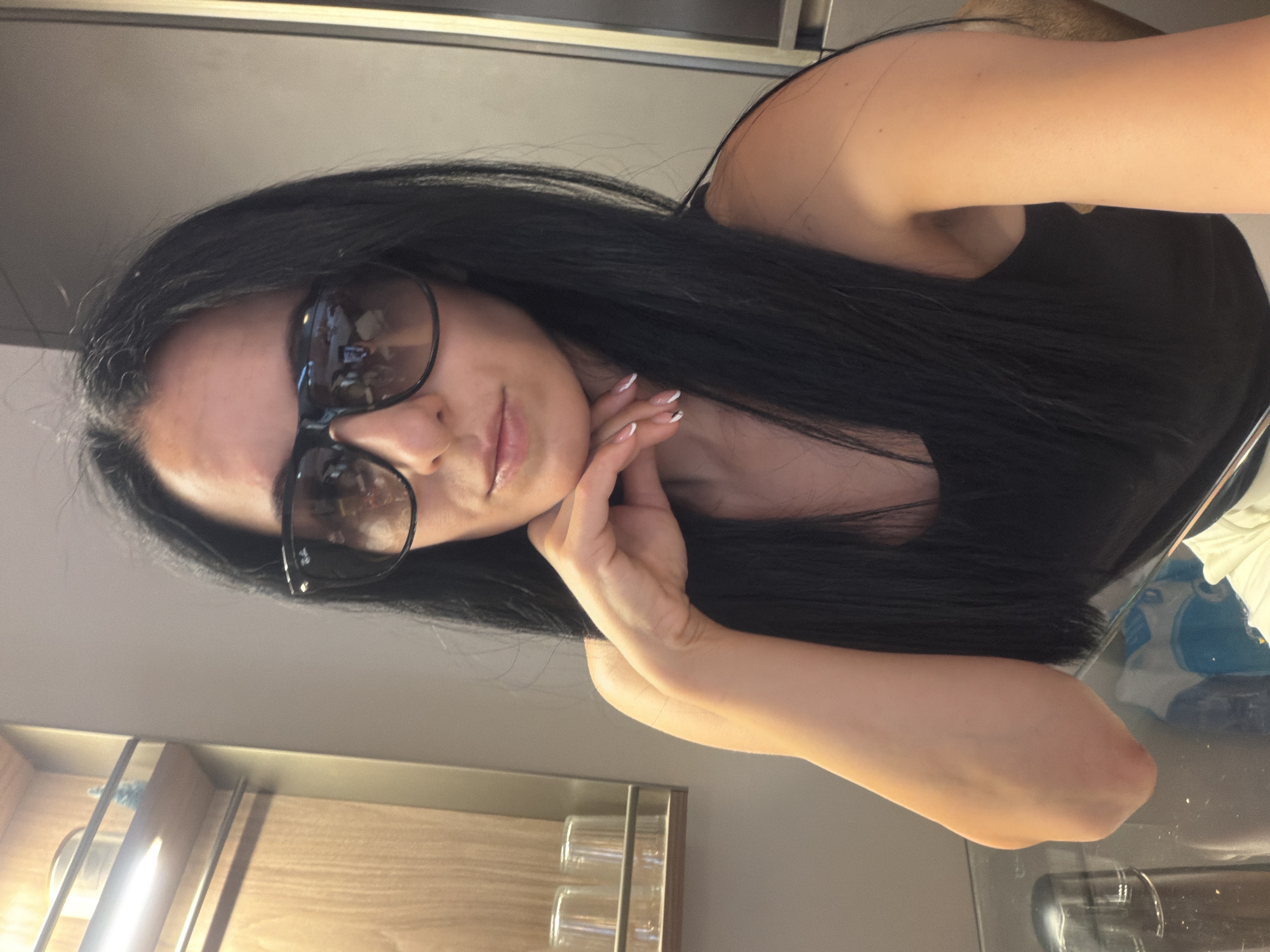 A medium shot captures a woman with long, dark hair and sunglasses, leaning her chin on her right hand. She wears a black tank top and white pants. The background features a wooden shelf with items on it, a grey wall, and a glass table. The image appears to be taken indoors, possibly in a hotel room or a similar setting, and no specific location within Buccinasco, Italy, is discernible from the visual information provided.