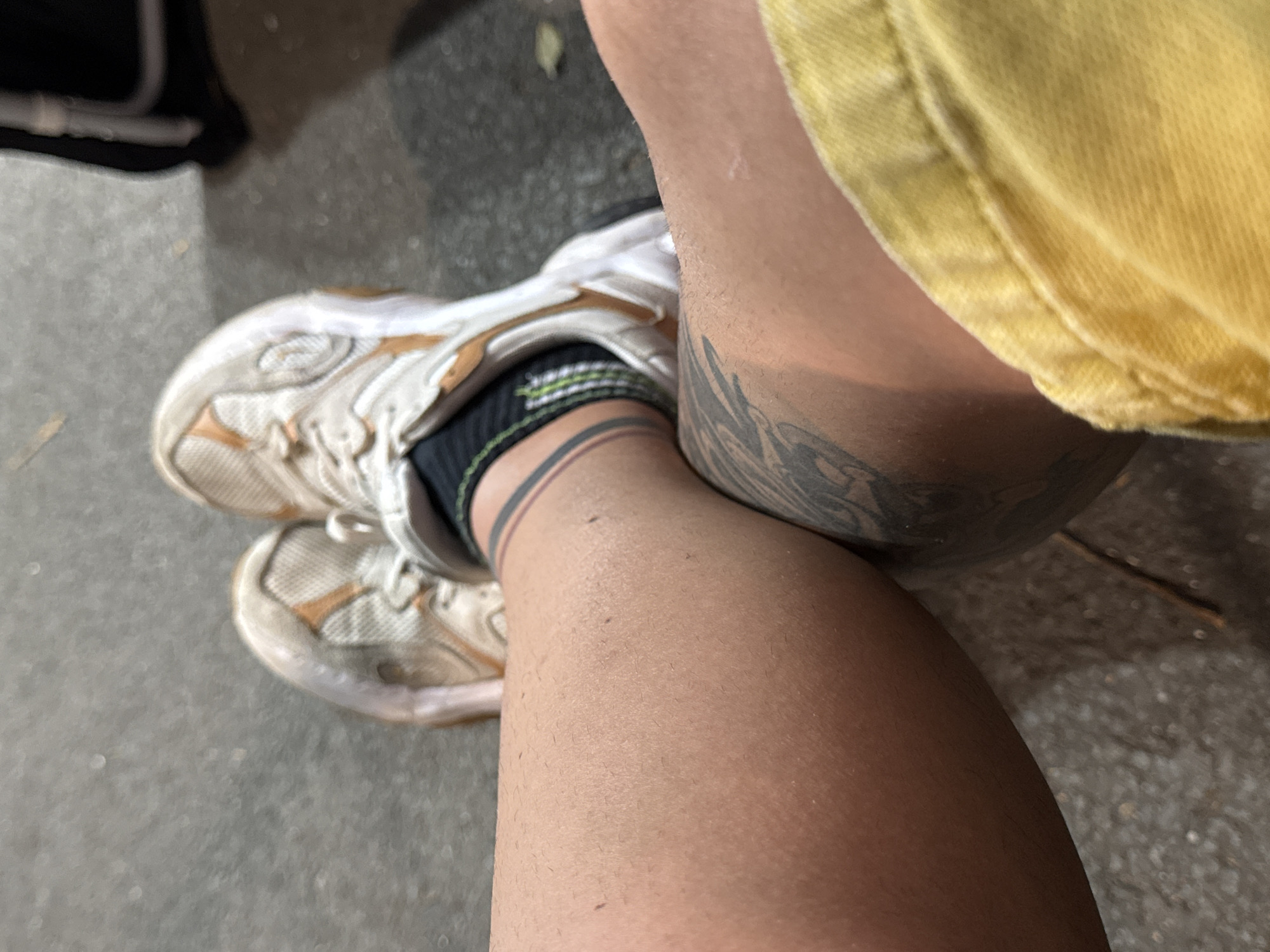 The image shows a close-up view of two people's legs. They are both wearing sneakers and socks. One person has a tattoo on their leg. The ground appears to be asphalt. The scene is outdoors, possibly in a public space like a park or sidewalk. There are no discernible landmarks to indicate the specific location in Burgas, Bulgaria.