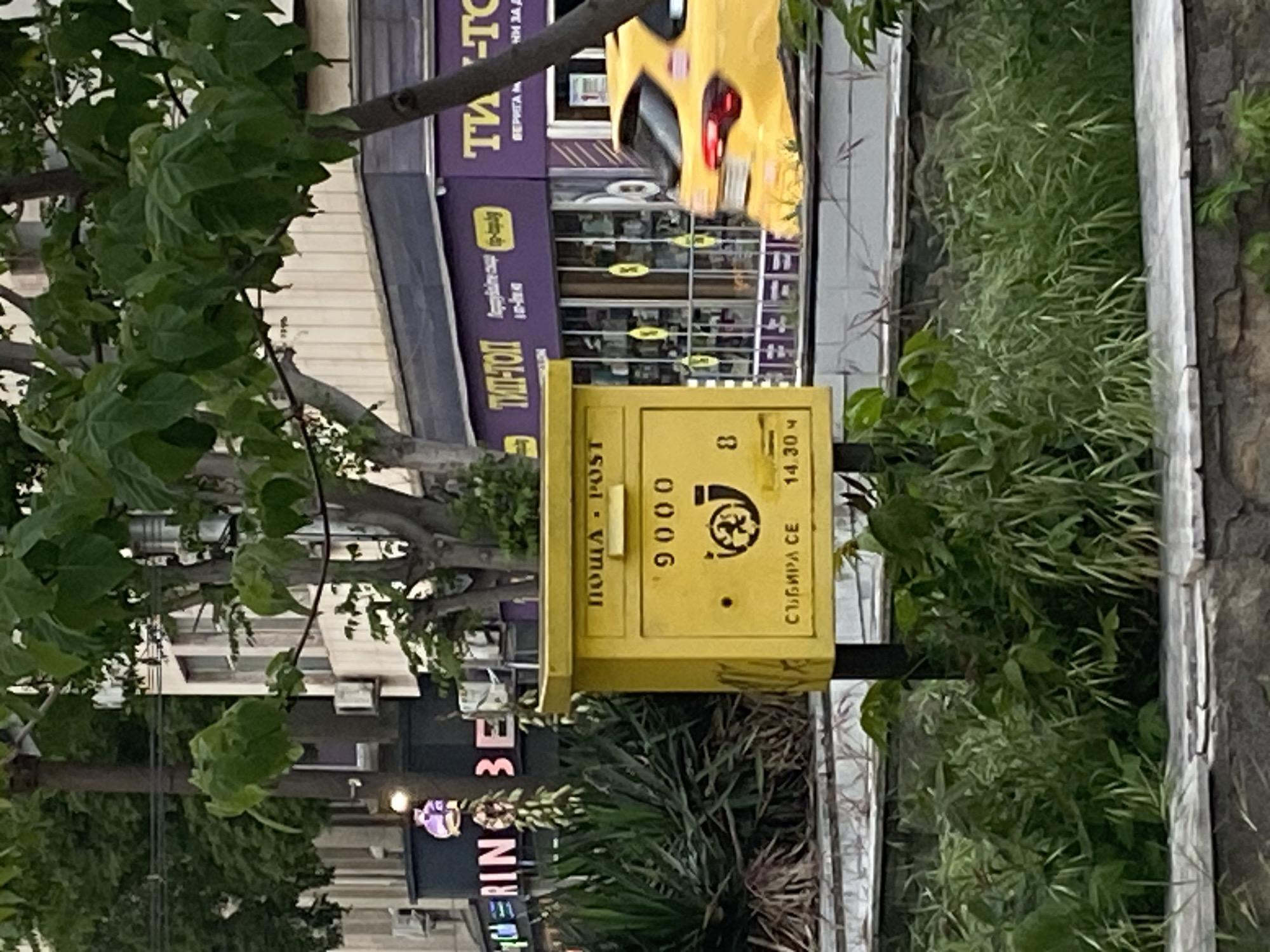The image shows a yellow post box in the city of Varna, Bulgaria. The post box is on a pole next to some greenery. It has the words "ПОЩА - POST" printed on it along with the number "9000" and a symbol with a picture of a man. The text "СЪБИРА СЕ" and "14.30 ч" are also on the box. The background includes buildings, foliage, and a glimpse of a yellow car.