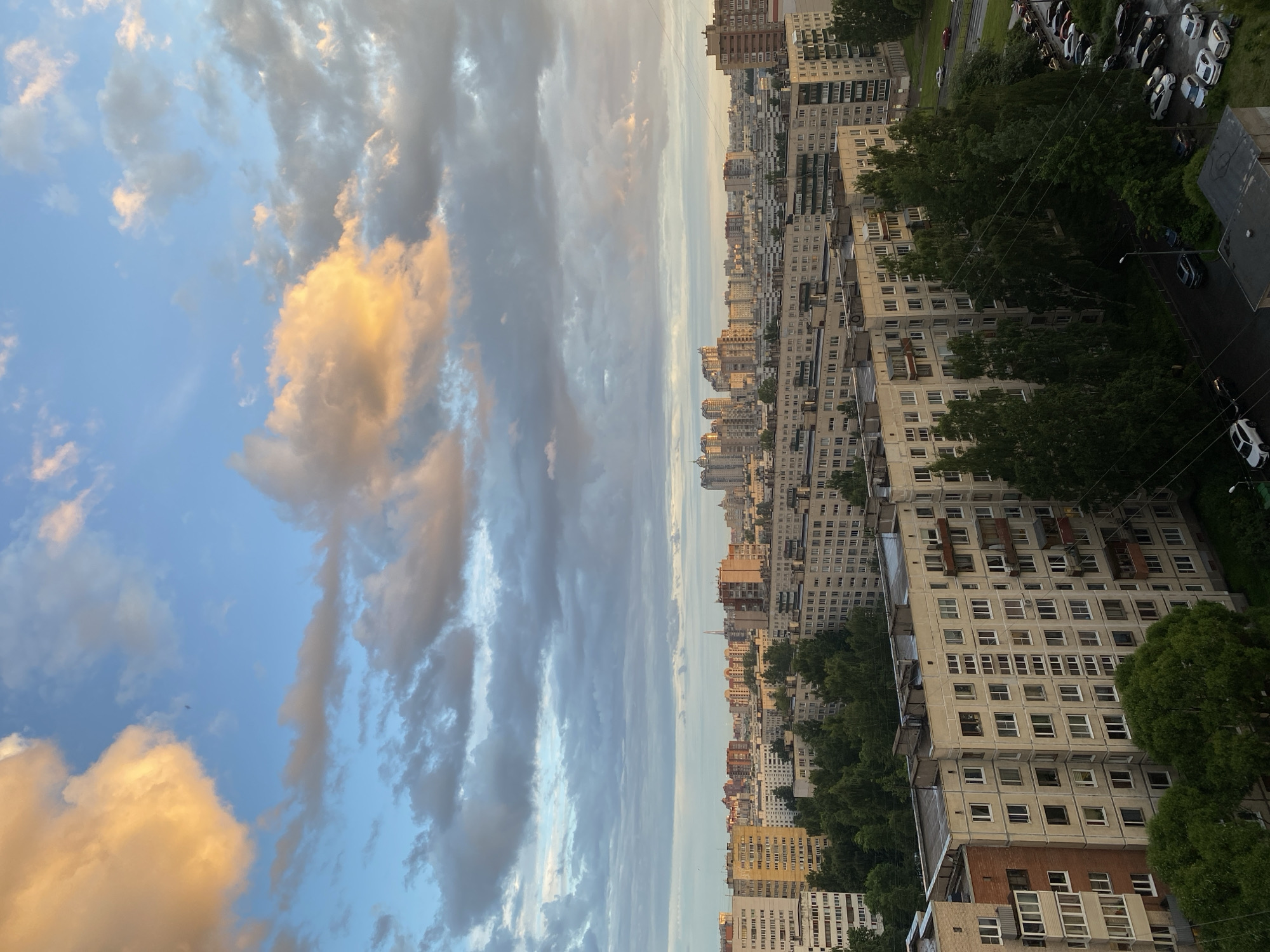 This is a vertical, full-shot photograph taken from a high vantage point, likely an apartment building, overlooking a city skyline. The scene is dominated by a vast expanse of sky filled with dramatic clouds. On the left side of the frame, the sky is a bright, clear blue, interspersed with patches of soft, golden-yellow clouds illuminated by the sun. As the eye moves towards the right, the clouds become denser, darker grays, and appear textured, suggesting an overcast or developing weather system.
The right side of the image features a dense urban landscape composed of numerous apartment buildings. These buildings are varied in height and architectural style, with many appearing to be older, monolithic Soviet-era structures characterized by repetitive window patterns and neutral-colored facades. Some buildings are clad in light beige or grey, while others have reddish-brown brick accents. Trees are interspersed throughout the urban landscape, some growing tall enough to partially obscure the buildings, and others forming a dense green canopy in the foreground. A street is visible with parked cars, mostly white and silver, suggesting a residential area. Power lines are also visible crisscrossing the scene.
The lighting suggests it is either sunrise or sunset, given the warm glow on some of the clouds and the long shadows that would be cast. The overall mood is somewhat dramatic due to the contrasting sky conditions. There is no visible text in the image. The scene depicts a typical urban environment under a dynamic sky.