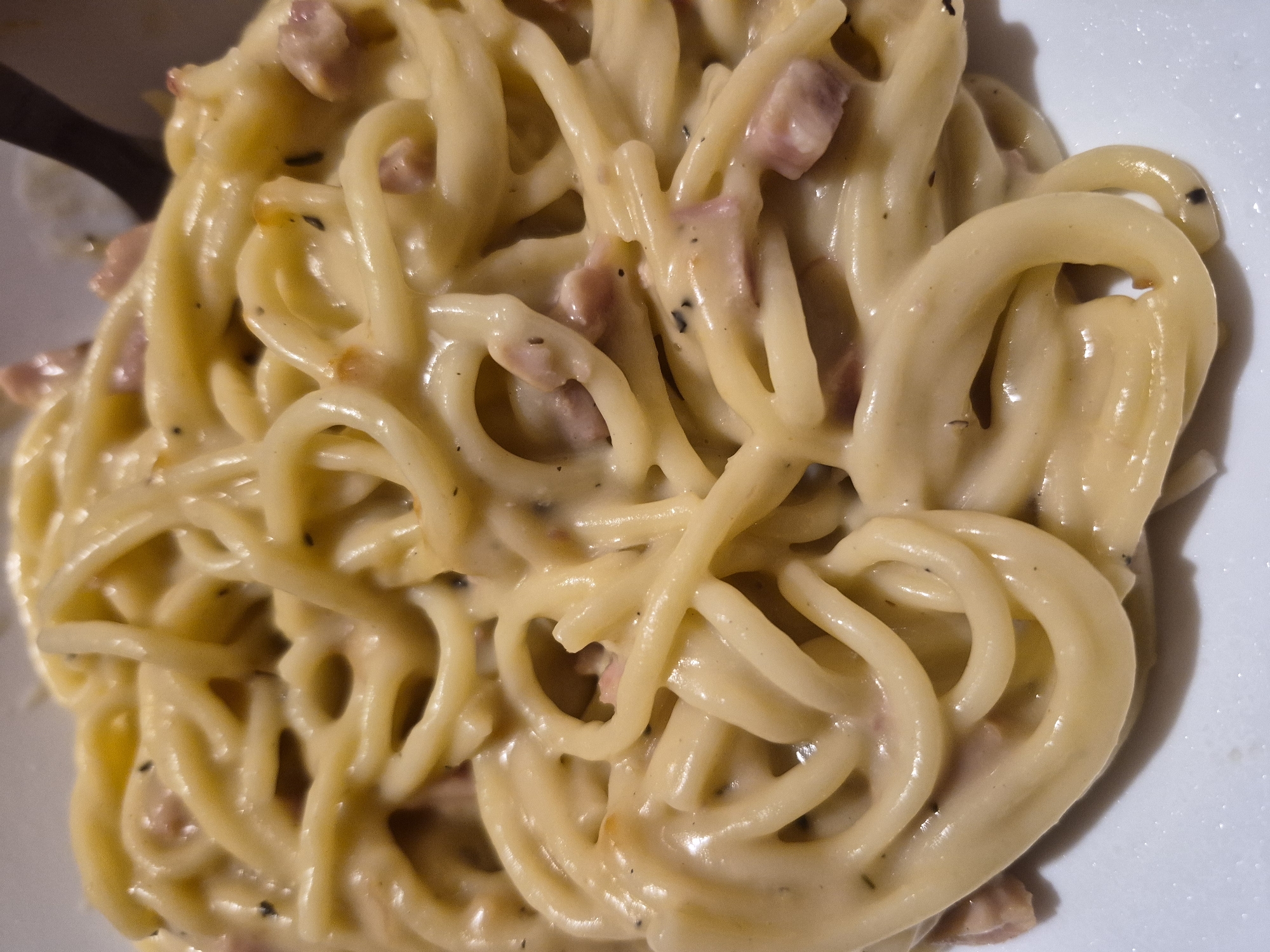 The image displays a close-up, overhead view of a plate of spaghetti carbonara. The pasta is coated in a creamy, pale yellow sauce, with visible pieces of diced pancetta or bacon interspersed throughout. Black specks, likely pepper, are also present in the sauce. The spaghetti strands are loosely coiled, filling the frame. A dark utensil, possibly a fork, is partially visible in the upper right corner. The background is out of focus and appears to be a white surface, likely the plate. The scene is indoors, with soft lighting that highlights the texture of the pasta and sauce. No people are visible in the image. The specific location in Sandwell, United Kingdom, cannot be determined from the visual information provided.