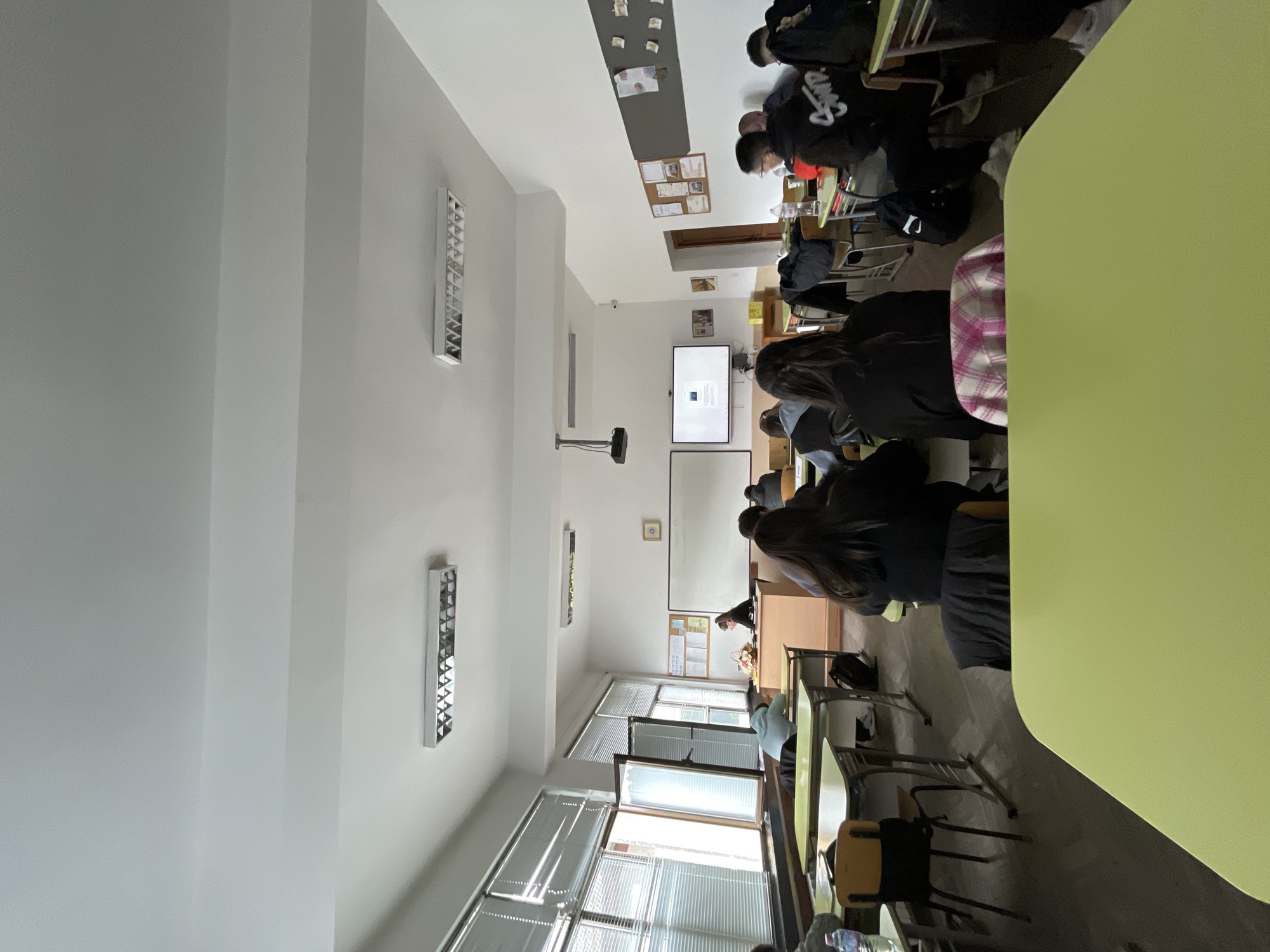 This image depicts an indoor classroom or meeting room in Zlatograd, Bulgaria, oriented vertically. The room features white walls and ceiling, with several long fluorescent light fixtures providing illumination. Large windows on the left side are covered with horizontal blinds, allowing daylight to enter.
Several individuals, mostly young adults or students, are seated at wooden desks with chairs, arranged in rows. Most are visible from behind, facing the front of the room. One person in the foreground wears a plaid shirt. In the background, what appears to be a teacher or presenter is positioned near the front.
At the front of the room, a projector is mounted on the ceiling, casting an image onto a white projection screen. To the right of this screen, a flat-screen television is also displaying content, which appears to be a document or presentation with paragraphs of text. While text is visible on both screens, it is too distant and blurry to be deciphered. A bulletin board with various papers and notes is mounted on the wall.
The scene suggests a class, lecture, or presentation is underway, with attendees seated and seemingly focused on the displayed content. A light green tabletop is visible in the bottom right corner, suggesting another desk or a counter close to the viewer. On one student's black hoodie, a partial logo or text resembling "SCMP" can be seen. The general lighting suggests it is daytime.