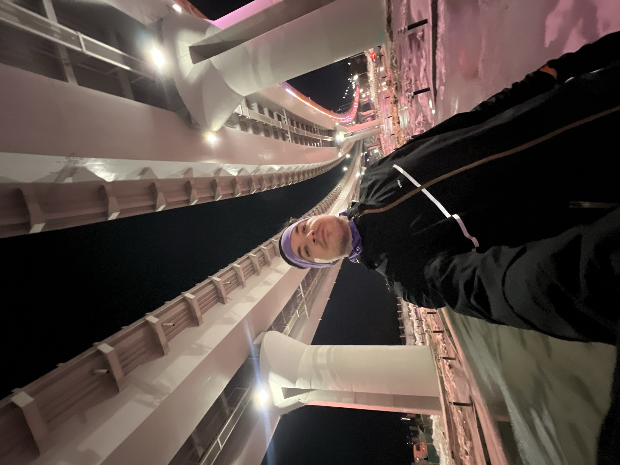 The image displays an adult male captured in a selfie at night, positioned directly underneath a multi-level elevated highway structure in Kazan, Russia. This architectural style, featuring robust concrete pillars and girders illuminated with bright white and distinct pink/purple artificial lighting, is characteristic of Kazan's modern road infrastructure, such as the interchanges near the Millennium Bridge or along the Kazan Ring Road.
The male subject, of fair complexion, occupies the lower-left to central portion of the frame, looking directly towards the camera with a neutral expression. He wears a black long-sleeved jacket adorned with a single diagonal white stripe and a vertical brown stripe on the wearer's left side. A purple headband or neck gaiter is worn across his forehead and ears. A dark glove is visible on his right hand, extended towards the camera, indicating he is holding the device.
The environment is illuminated by artificial lights emanating from the elevated structure and other distant sources. The ground beneath the overpass is paved and appears wet, reflecting the light, with visible patches of snow and ice, indicating winter conditions. A series of dark, short bollards or posts line the edge of a paved area on the right. In the background, sections of the massive elevated highway curve away, displaying prominent pink and white accent lighting. Faint structures, possibly industrial or construction-related, are discernible in the far-left background beyond a snow-covered bank. No other individuals are visible, and the subject's interaction is solely with the camera.