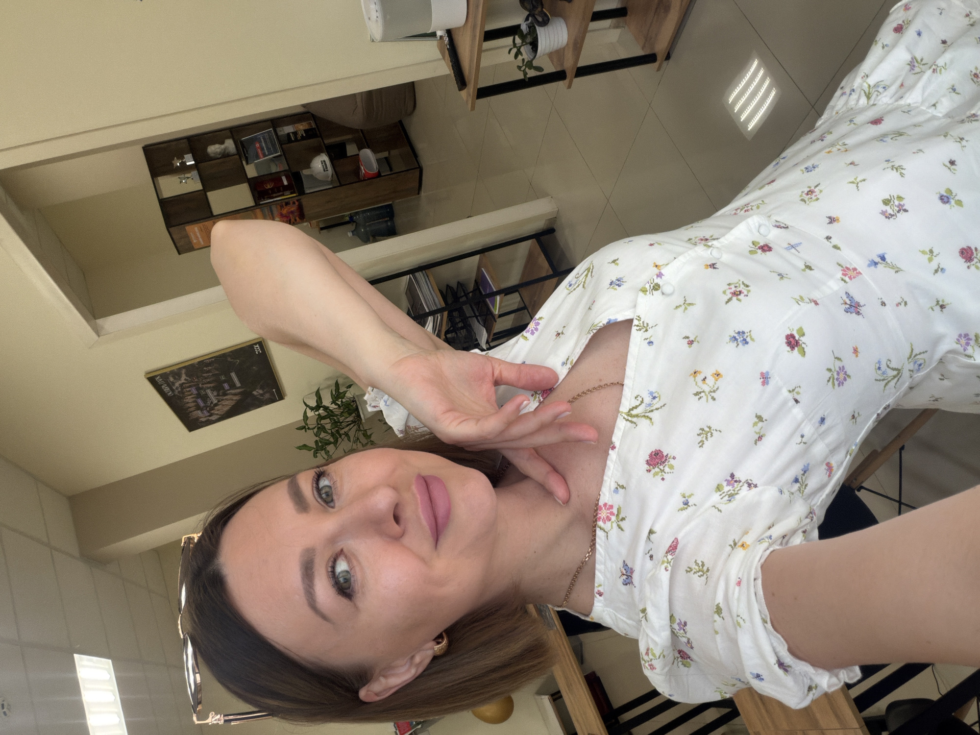 A woman is posing for a selfie in what appears to be an indoor setting. She is wearing a white dress with a floral pattern and a gold necklace. The background includes shelves with various items, a picture frame on the wall, and a glimpse of a window. The precise location within Rostov-on-Don, Russia, cannot be determined from the provided image.