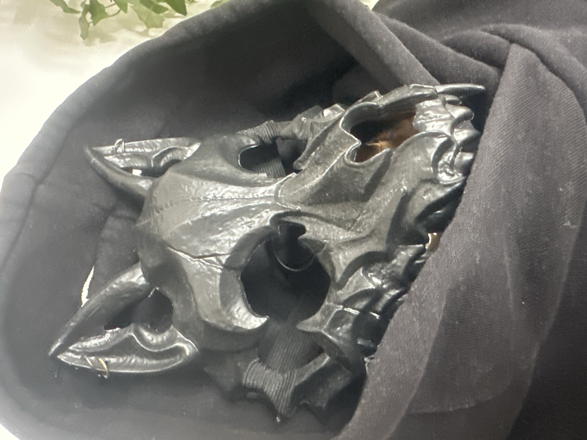 A black, detailed, dragon-like mask is nestled within the folds of a black hooded garment. The mask, made of what appears to be a hard material, features sharp angles, protruding horns, and an open mouth revealing a glimpse of what might be a wooden or amber-colored inner lining. Two small, silver-colored rings are attached to the outer edges of the mask, likely for attaching straps. The fabric of the hood is soft and textured, with visible stitching and subtle creases, suggesting it is made of cotton or a similar material. The lighting in the image is soft and diffused, casting gentle shadows and highlighting the mask's intricate design. In the background, out of focus, are hints of greenery, possibly leaves or branches, and a bright, light-colored surface, which could be a wall or table. The overall impression is one of a costume piece or prop, possibly prepared for an event or simply stored. The image focuses closely on the mask and its immediate surroundings, giving a sense of depth and texture.