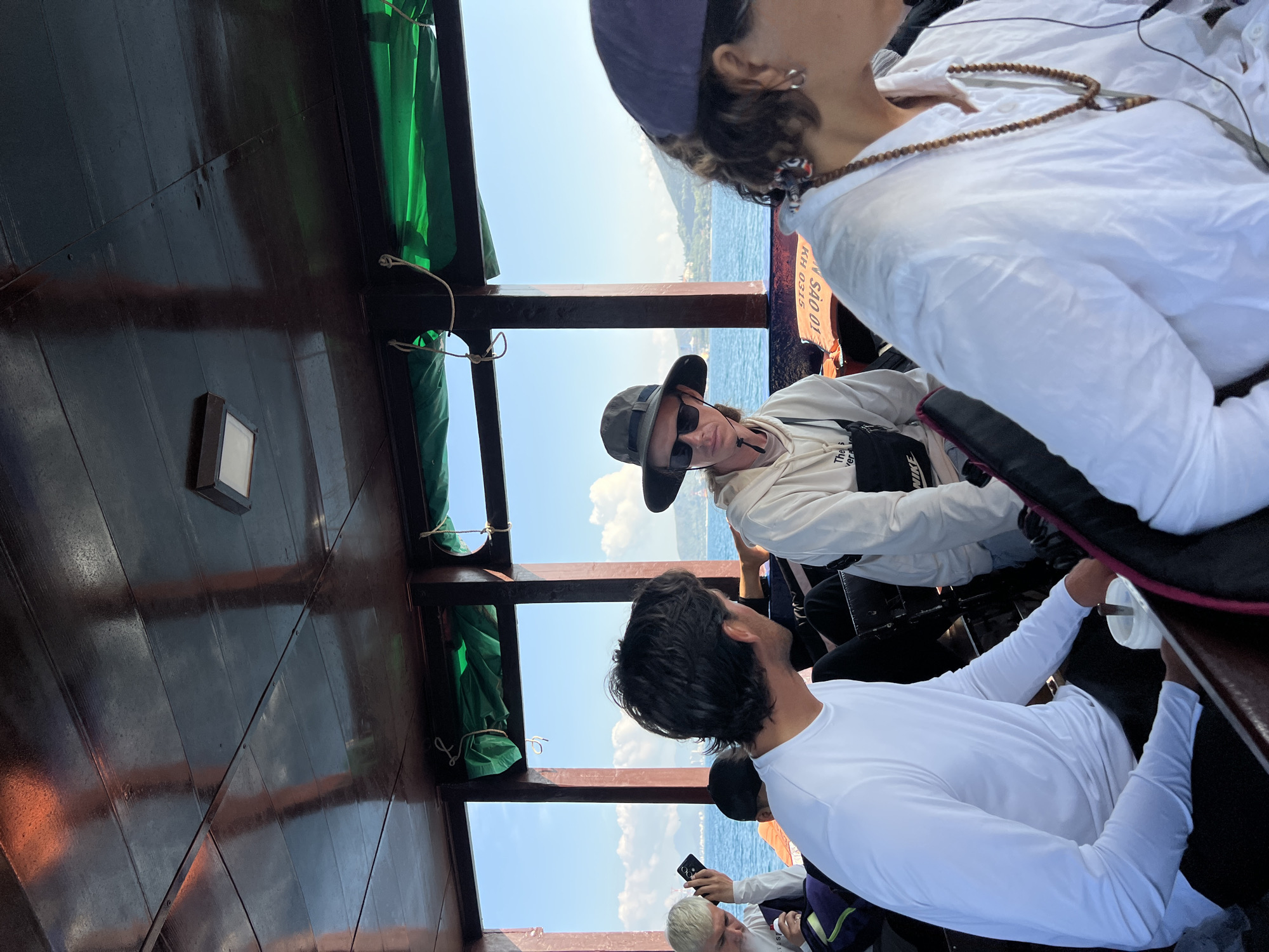 The image appears to be taken from inside a boat, likely on the water. The setting suggests a coastal or marine environment.
Several individuals are visible. One person in the center wears a hat and sunglasses and a white top. Another individual wearing a white shirt with long sleeves and dark hair is in the foreground. Others are visible in the background, seemingly seated. One appears to be taking a picture with a phone.
The structure around the people consists of dark wood, likely the boat's interior. Green curtains are visible towards the upper left portion. The sky and water are visible through openings or windows.
The boat is possibly situated in or near Bac Nha Trang Ward, Vietnam, given the presence of Vietnamese text on the boat's exterior.