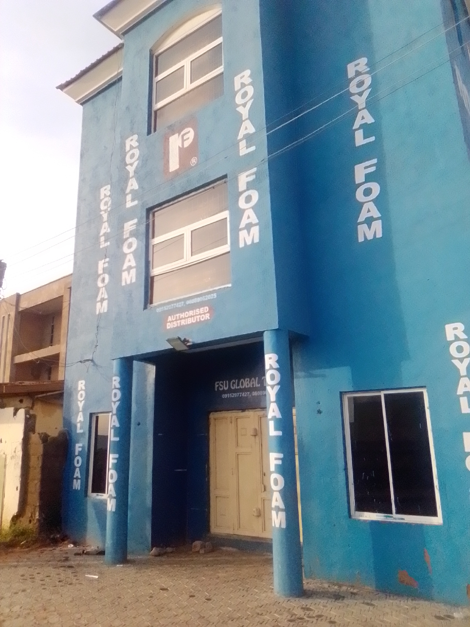 The image shows the exterior of a blue building in Pantame, Nigeria, with the words "ROYAL FOAM" painted vertically in white on its facade multiple times. The building has several windows, some with white frames and dark glass. A smaller building, appearing to be an attached structure or a separate building adjacent to it, is visible on the left. The ground in front of the building is paved with interlocking bricks. There are some wires running horizontally across the building. Text below a window on the building reads "FSU GLOBAL" and a phone number, with "AUTHORISED DISTRIBUTOR" below it. The sky is overcast.