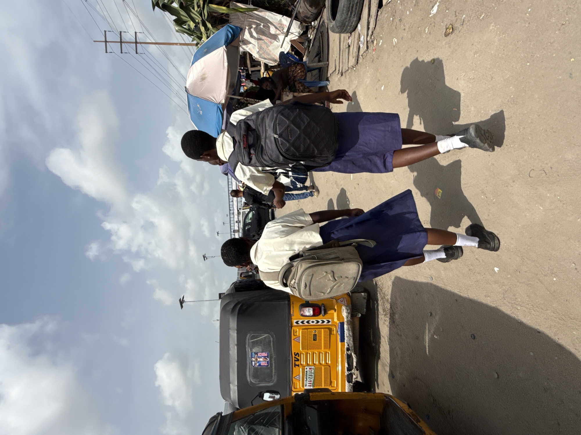 Two students in school uniforms walk along a street in Lekki, Nigeria. The students wear white shirts and blue skirts, with backpacks and white socks. A yellow and black auto rickshaw, a common mode of transportation in Nigeria, is parked nearby. In the background, there is a street vendor with a blue and white umbrella covering their goods. Utility poles and overhead wires are visible against a sky with scattered clouds. The image captures a typical street scene with children in uniform going to or from school.