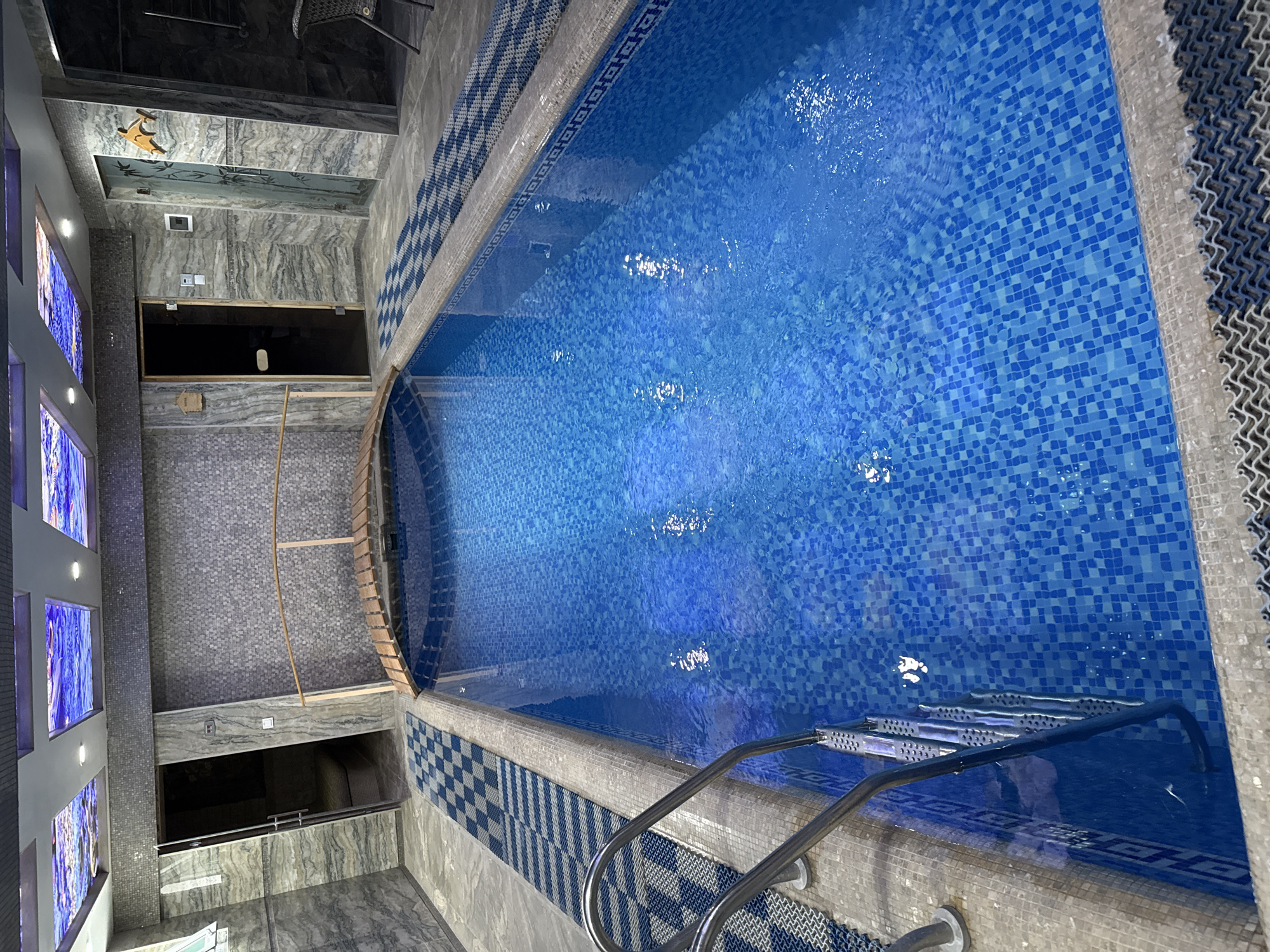 The image shows an indoor swimming pool in what appears to be a private residence or a small hotel. The pool is filled with clear blue water and lined with blue mosaic tiles. A metal ladder provides access to the pool. The pool's perimeter is decorated with patterned tiles in blue, white, and gray. The surrounding walls and floor are tiled in gray. There are some lighting fixtures. The location is potentially in Tashkent, Uzbekistan. There are no people visible in the image.