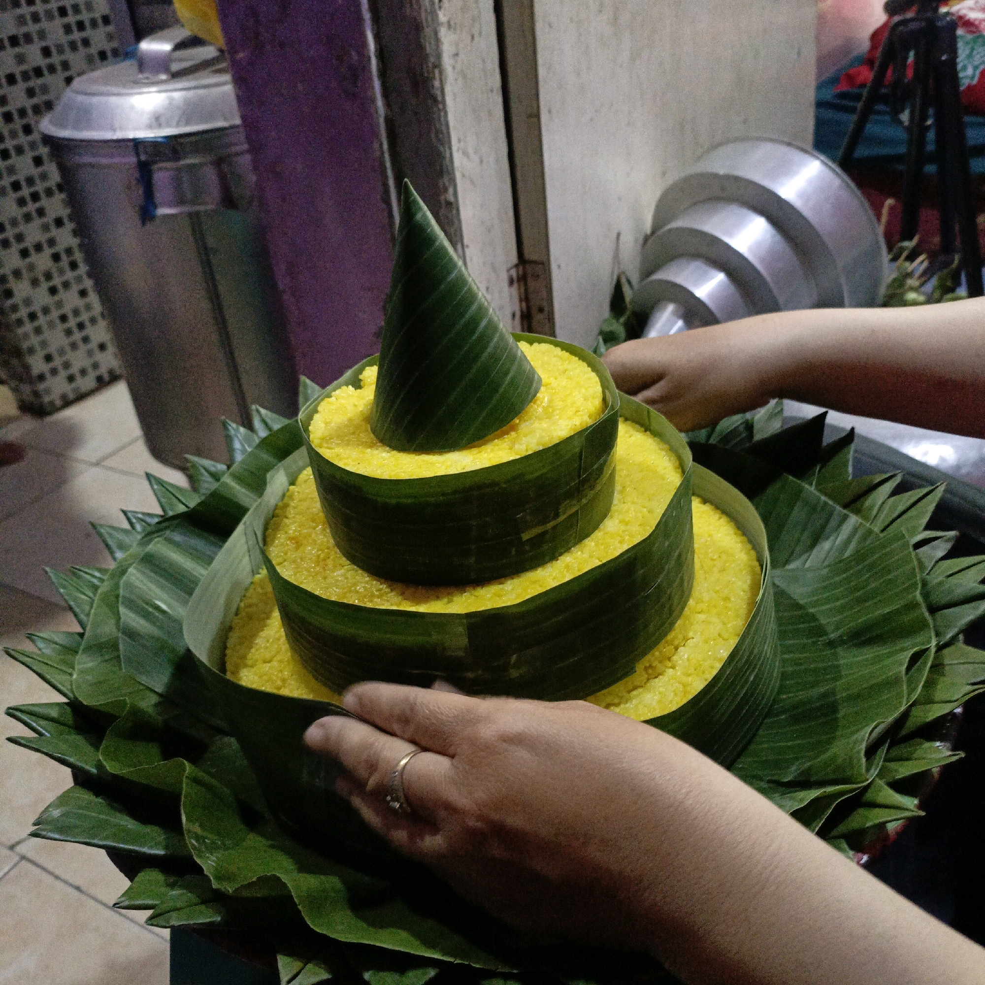 The image shows a three-tiered, conical cake made of yellow rice, decorated with green banana leaves. The cake is placed on a large platter also lined with banana leaves. Two people's hands are visible, one adjusting the banana leaf decorations at the base of the cake, and another's hand is seen on the right side of the frame. In the background, a silver cylindrical container with a handle is partially visible on the left, and a stack of nested aluminum bowls is on the right. The scene appears to be indoors, possibly in a kitchen or preparation area, with a tiled floor and a light-colored wall. The lighting suggests it is daytime, but the setting is somewhat dim. This presentation is typical of a tumpeng, a cone-shaped rice dish often served at celebrations in Indonesia, including in the city of Salatiga.