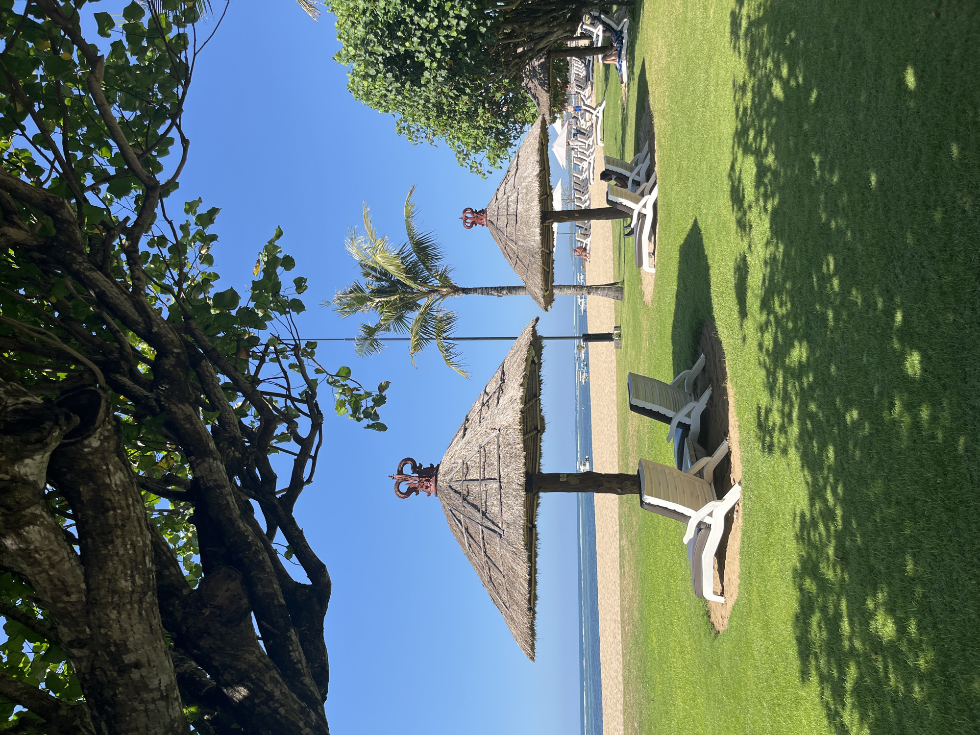 The image depicts a serene beach resort scene in Kuta Selatan, Indonesia, under a clear blue sky. In the foreground, lush green grass is dappled with sunlight filtering through the canopy of a large tree. Two white lounge chairs with grey cushions are positioned on the grass, partially shaded by a thatched umbrella with a distinctive red, crown-like ornament on top. Beyond the chairs, a sandy beach meets the calm, blue ocean, where a few boats are visible in the distance. A solitary palm tree stands tall behind the umbrella, adding to the tropical ambiance. Further back, more thatched umbrellas and lounge chairs are arranged on the sand, some occupied by individuals relaxing. A line of trees borders the far side of the beach. The overall impression is one of tranquility and a tropical vacation setting.