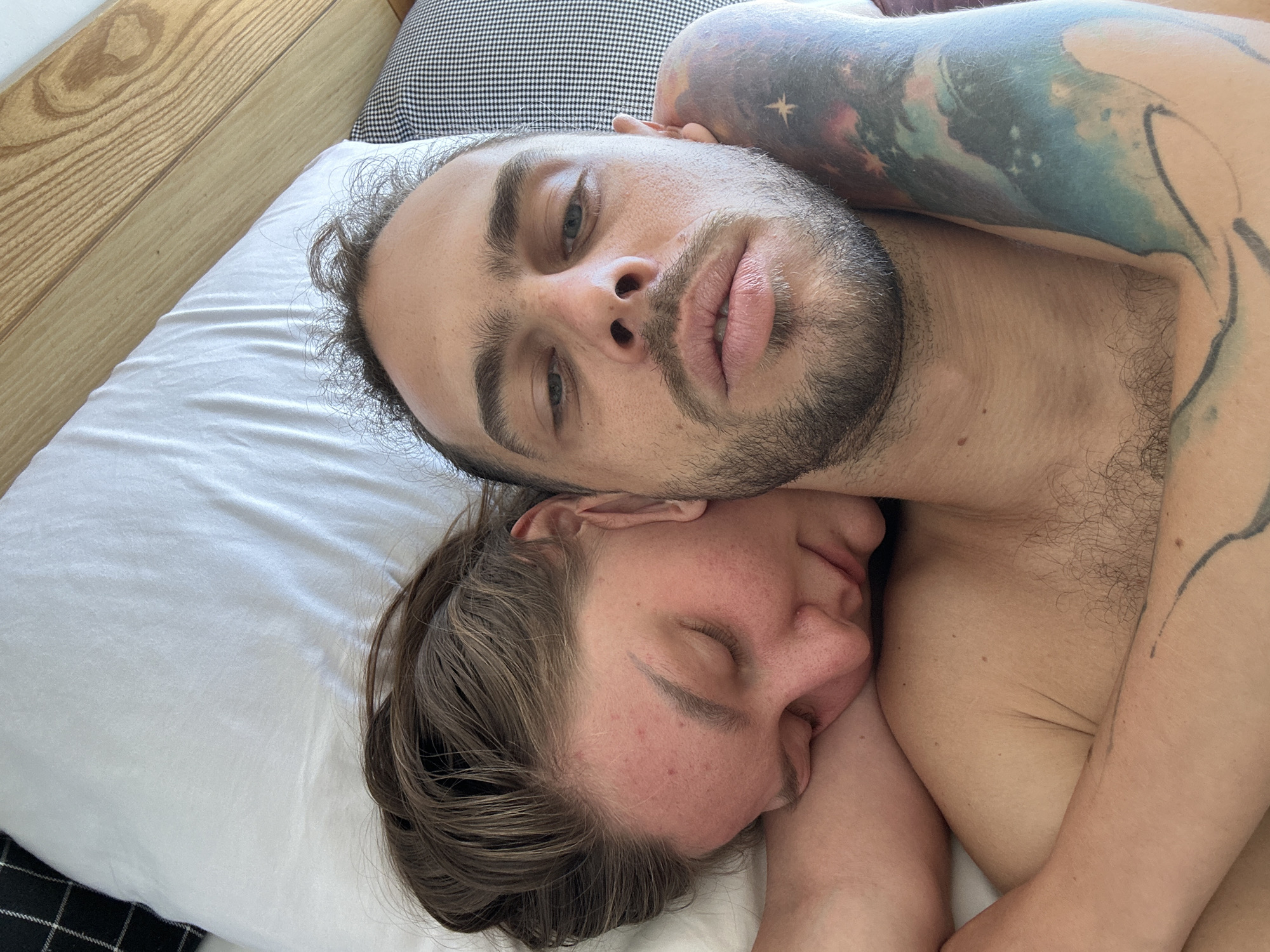 The image shows a close-up, overhead view of two people lying in bed, embracing. The person on top, facing the camera, has light skin, short brown hair, and stubble. They are looking at the camera with their eyes open, and their lips are slightly parted. A large, colorful tattoo of a galaxy is visible on their left arm and shoulder. The person below, with their head resting on the shoulder of the person above, has their eyes closed and appears to be asleep. They have light skin and brown hair. The bedding is white, and a pillow with a black and white checkered pattern is visible in the background. The scene is indoors, likely a bedroom. There is no identifiable information in the image that places the scene in the city of Đà Nẵng, Vietnam.
