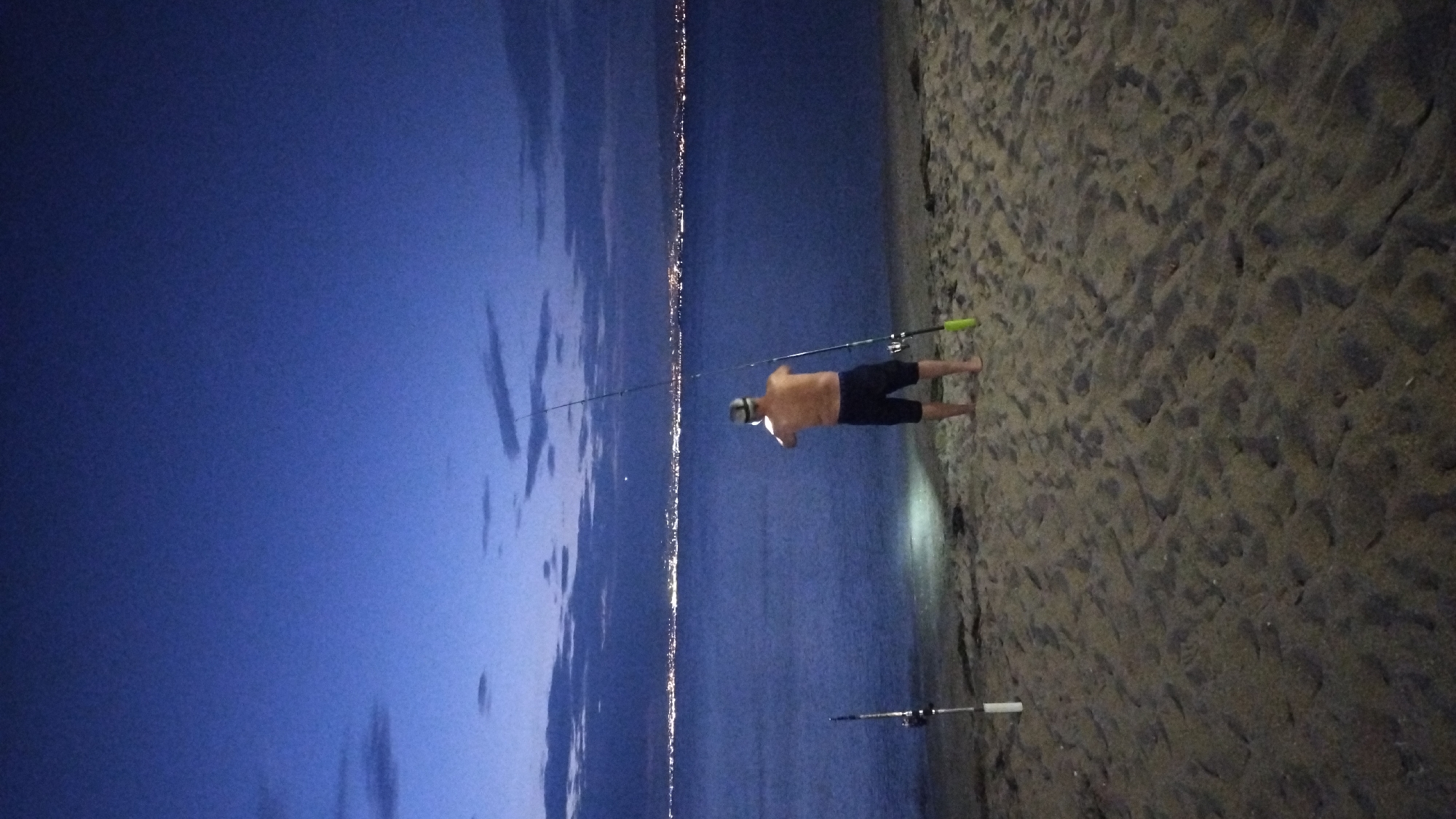 A lone, shirtless man stands on a sandy beach in Perea, Greece, facing a body of water as dusk settles. He is holding a fishing rod, and two other fishing rods are planted in the sand beside him. The water stretches to the horizon, where a city skyline is illuminated by countless lights. The sky above is a deep blue, with scattered clouds. The man is wearing dark shorts and a cap. A bright light shines from the sand at his feet, illuminating the area around him.
