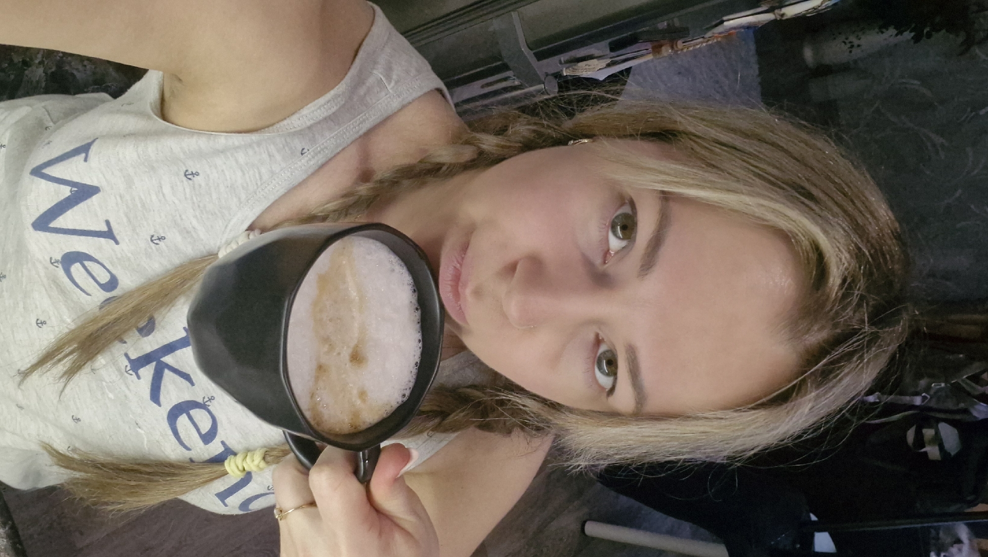 A fair-skinned woman with blonde hair in pigtails is holding a black mug filled with a frothy, light brown beverage. She is wearing a light grey tank top with blue lettering and small anchor designs. She has light-colored eyes and is looking at the camera. She has a gold ring on her hand. The environment appears to be indoors, with a dark-colored surface visible in the background. The location is unidentifiable as Otchor, Russia based on the visual information.
