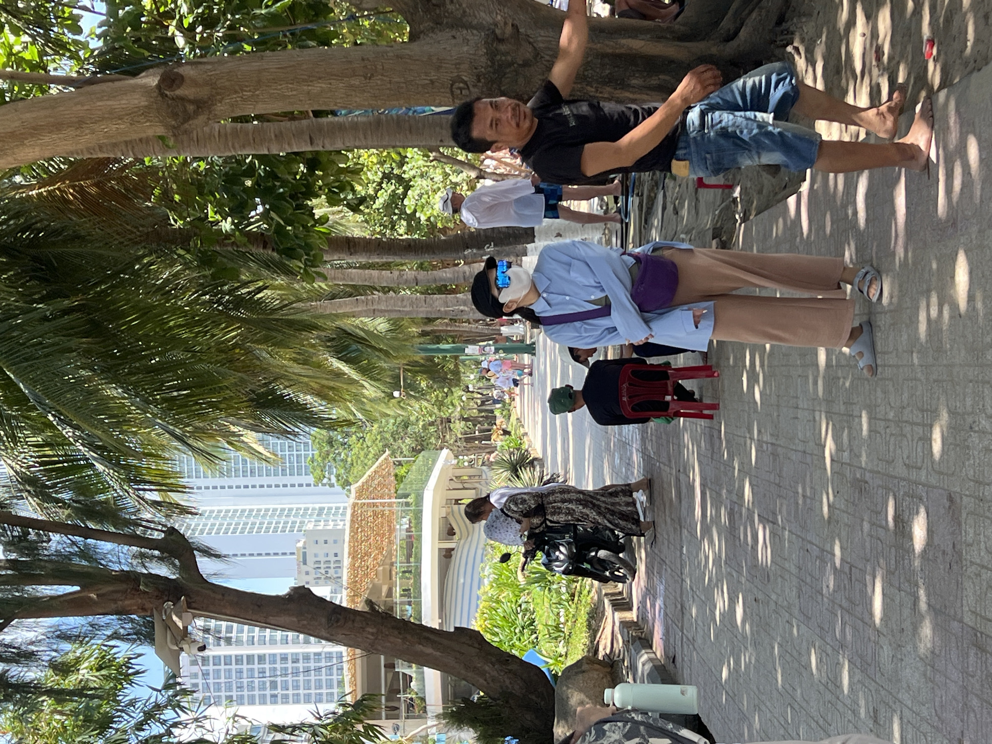 The image shows a street scene in Nha Trang Ward, Vietnam. A mature tree dominates the left foreground, its branches extending across the frame. Tall buildings are visible in the background. A sidewalk with interlocking pavers runs through the right half of the image. Several people are present: a man in a black shirt and blue shorts is standing near a tree; a woman with a face mask and a purple bag is walking on the sidewalk; and another woman is near a motorcycle. A red plastic chair is also visible on the sidewalk. Other pedestrians are further down the street.