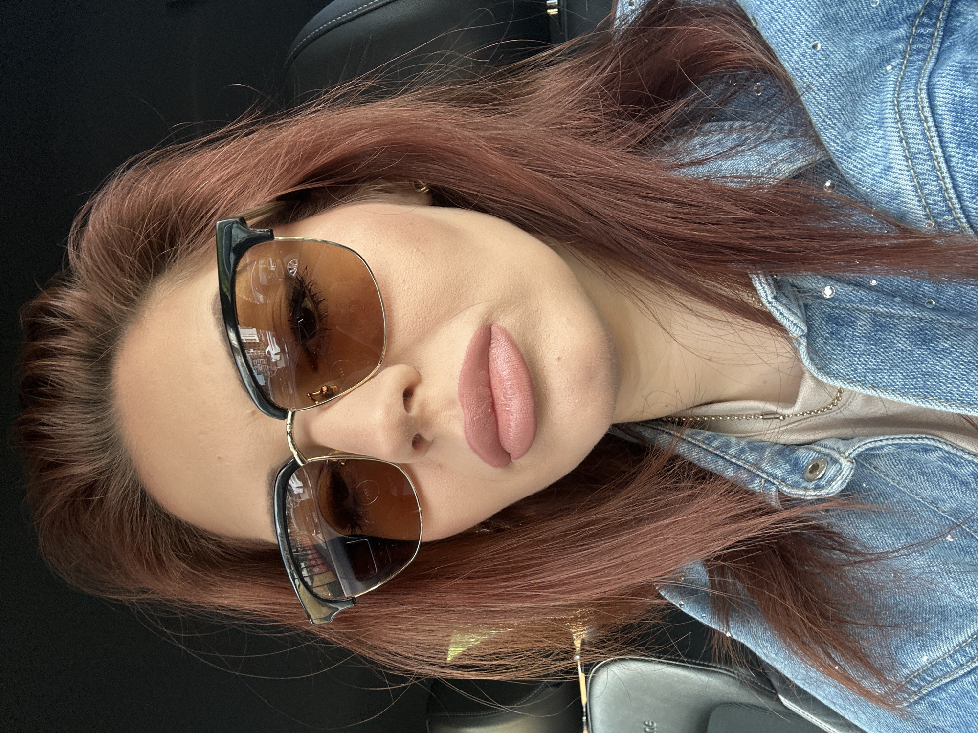 A woman with reddish-brown hair is lying down in a car. She is wearing sunglasses with black and gold frames and brown lenses. She has nude lipstick on. She is wearing a light blue denim jacket with silver embellishments and a beige top. The location is unknown and there is no information to indicate that the scene is in Sofia, Bulgaria.
