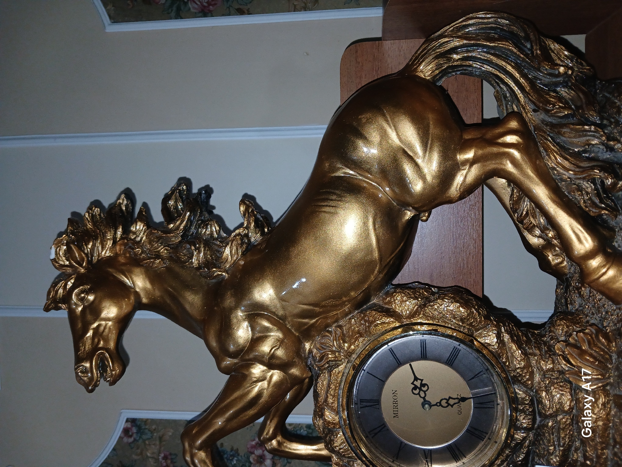 The image shows a close-up of a decorative gold-colored clock featuring a rearing horse sculpture. The horse is depicted in motion, with its mane and tail flowing. The clock face is round with Roman numerals and has "MIRRON" and "QUARTZ" visible on it. The clock is part of a larger decorative piece that is golden and appears to be made of resin or metal. The background consists of a wall with floral wallpaper at the bottom and a lighter-colored wall with molding above. A wooden shelf is visible on the right side. The lighting suggests an indoor setting, possibly in the evening or with artificial light. The image has a watermark on the right side that reads "Galaxy A17".
