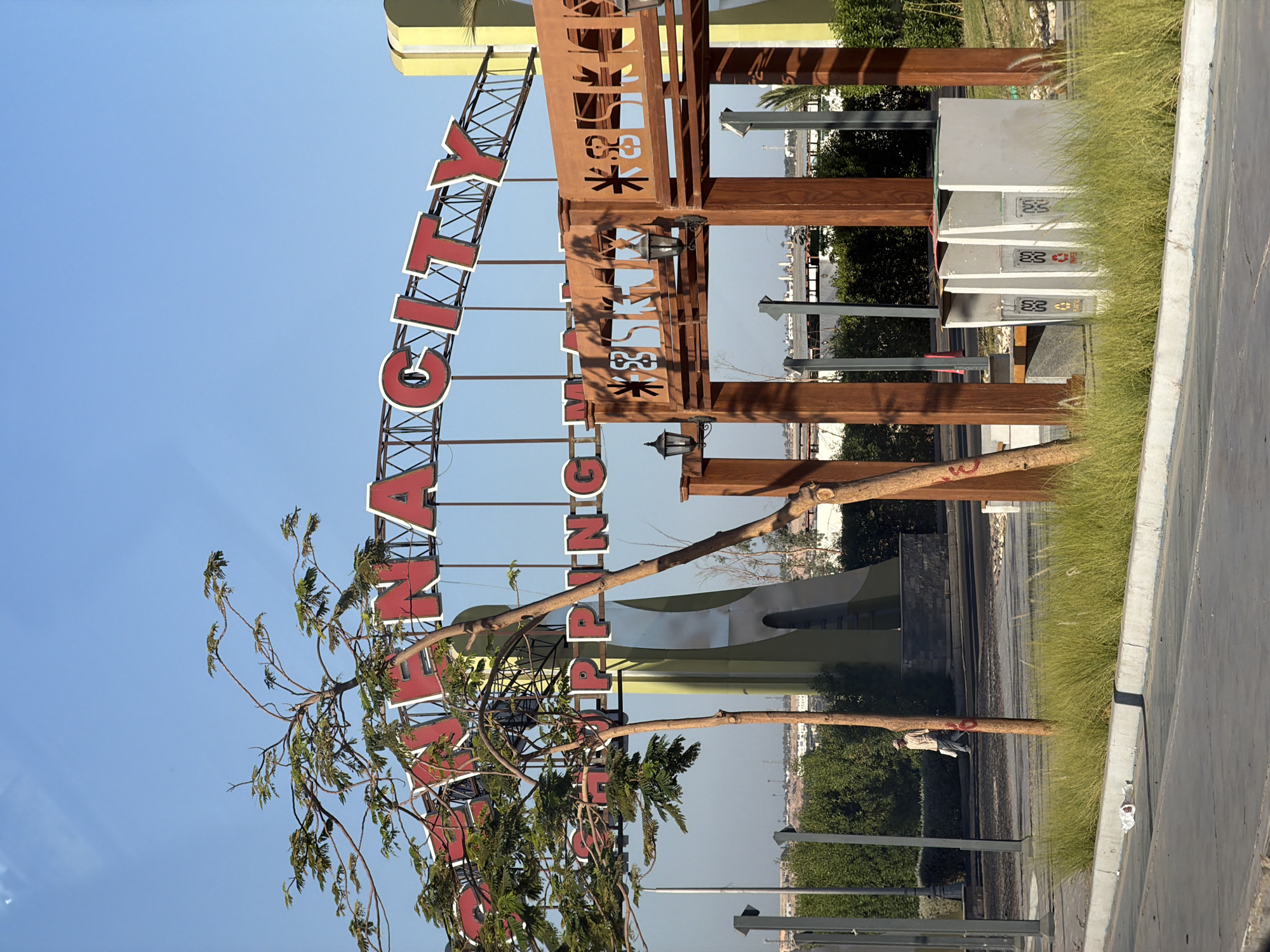 The image depicts the entrance to a shopping mall in Sharm El Sheikh, Egypt, with a large sign overhead that reads "Shopping City." The sign is made of red letters with white outlines, and it is mounted on a metal truss structure. In front of the sign, there are two trees, one taller and thinner than the other, with green foliage.
Below the "Shopping City" sign, there is another sign with the words "SHOPPING MA." This sign is also made of red letters with white outlines, and it is mounted on a wooden structure. The wooden structure appears to be an archway with decorative cutouts and two black lanterns.
In the background, there is a tall, slender building with a light green and white color scheme, resembling a tower. Beyond the buildings and trees, a cityscape with buildings and possibly hills can be seen under a clear blue sky.
In the foreground, there is a paved area, possibly a road or walkway, with a gray concrete curb and a strip of green grass. A single person wearing a light-colored shirt and a hat is walking on the paved area in the middle ground. There are also some trash bins visible near the wooden structure. The overall scene suggests a sunny day in an outdoor commercial area.