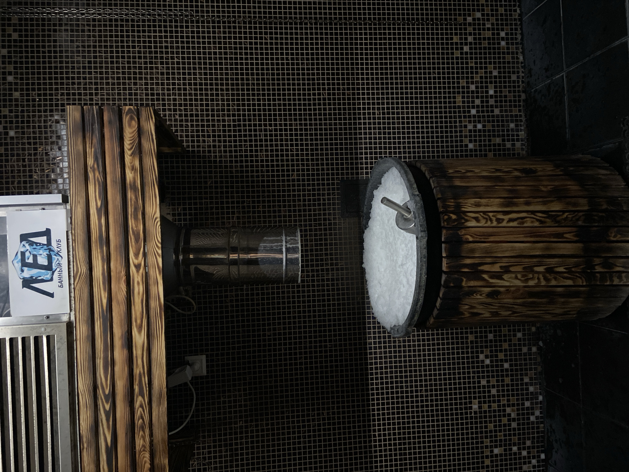 This image captures an indoor ice room or cooling area, likely within a bathhouse or spa, located in Kaliningrad, Russia. No people are present in the scene.
The primary subjects are a wooden bench, a metal ice-making machine, and a wooden barrel filled with crushed ice. The walls are covered in dark, small mosaic tiles forming a grid pattern, with some lighter, reflective tiles interspersed. On the left, a sturdy wooden bench with a dark, burnt-wood finish is positioned next to a stainless steel ice dispenser or machine. To the right, a rustic wooden barrel, also with a dark wood finish, holds a large quantity of white, crushed ice. A metal scoop is partially submerged in the ice, indicating it's meant for patrons to use.
The setup suggests the activity of cooling down after a hot treatment, such as a sauna or banya session. Guests would use the scoop to retrieve ice from the barrel, likely for rubbing on their bodies to stimulate circulation and refresh themselves.
Notable details include the rich textures of the dark wood and the mosaic tiles. The lighting is artificial, suitable for an indoor, specialized spa environment. A visible sign, partially cropped on the left, has Cyrillic text: "ЛЕД" (LED), meaning "Ice," and "БАННЫЙ КЛУБ" (BANNYY KLUB), meaning "Bath Club." This text confirms the room's purpose and the nature of the establishment.