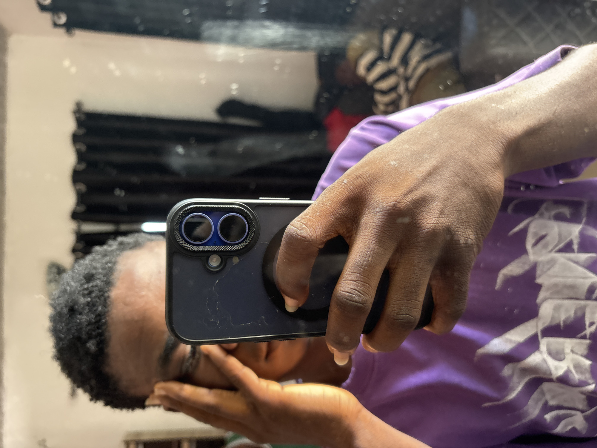 A person is taking a selfie in a mirror. The person is holding a dark-cased smartphone up to the mirror with their right hand. The person is wearing a purple t-shirt. The reflection shows the person's face, partially obscured by their left hand, and the reflection of the surroundings. The environment is indistinct, but a striped item is visible in the background. The location in the city of Ikot Ekpene, Nigeria, is not directly identifiable from the image.