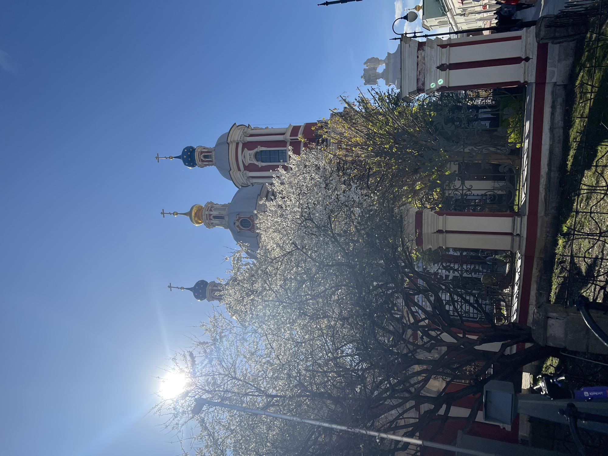 The image shows a low-angle shot of the Peter the Great High Monastery of the Moscow Patriarchate in Moscow, Russia. The architecture features a traditional Russian Orthodox style with multiple domes topped with crosses, painted in white, red, gray and gold. A blossoming tree with white flowers is prominently in the foreground, partially obscuring the lower portion of the monastery. The bright sun appears to be low in the sky, situated to the left. A fence and a street light can be seen to the right of the building. The sky is clear and blue.