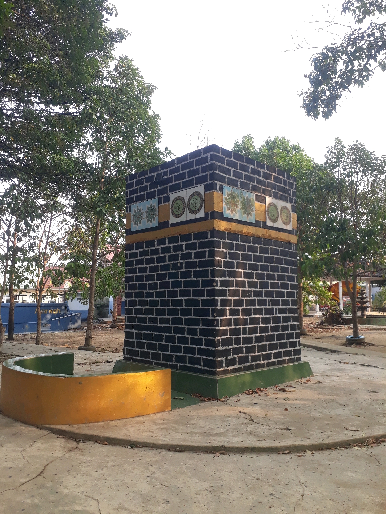 The image depicts a replica of the Kaaba, the holiest shrine in Islam, situated in a public space. This replica is constructed from black bricks with white mortar lines and is adorned with gold trim and decorative tiles featuring Arabic calligraphy and geometric patterns. The structure stands on a concrete platform, surrounded by a curved, golden barrier with a green interior. In the background, several trees with green foliage are visible, along with a clear sky. Further back, the roofs of buildings and other structures can be seen, suggesting an outdoor setting within a populated area. The overall environment appears to be well-maintained, with some fallen leaves scattered on the ground. Based on available information, this Kaaba replica is located in the city of Pangkalpinang, Indonesia, and serves as a religious landmark or exhibit. There are no people or animals visible in the image.