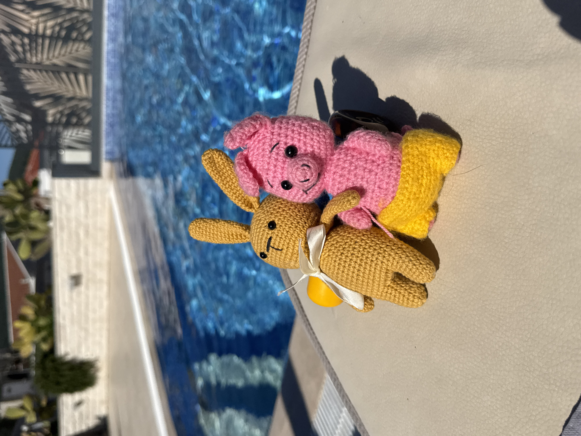 Image of a sunny outdoor scene showing a crocheted yellow rabbit and pink pig toy lying on a light-colored surface next to a pool in Đenovići, Montenegro. The water in the pool is clear and reflects the surrounding buildings and vegetation. The toys are the primary subjects of the photo. The rabbit has a white ribbon tied around its neck. The pig is positioned on top of the rabbit. A yellow item is beneath the pig.