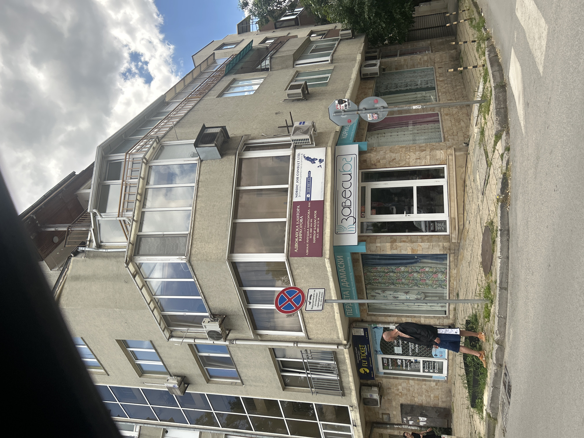 The media file shows a street view of a multi-story building in Varna, Bulgaria, on a bright, partly cloudy day. The building has a mix of commercial establishments on the ground floor and apartments or offices above. Visible businesses include a law office ("АДВОКАТСКА КАНТОРА КИРКОРОВА"), a Nordic Job Consult, and a curtain store ("ПЕРДЕТА | ДАМАСКИ" and "завесиба"). One of the ground floor entrances has a sign for "FLY TICKET" and a man in a black jacket and blue shorts standing near the doorway. A traffic sign indicating "No Stopping" is visible on a pole. The building's facade is a mix of stucco and stone cladding, with multiple windows and air conditioning units. A metal ladder runs up the side of the building. The street in the foreground has a pedestrian crossing. The overall scene depicts a typical urban street in a European city.