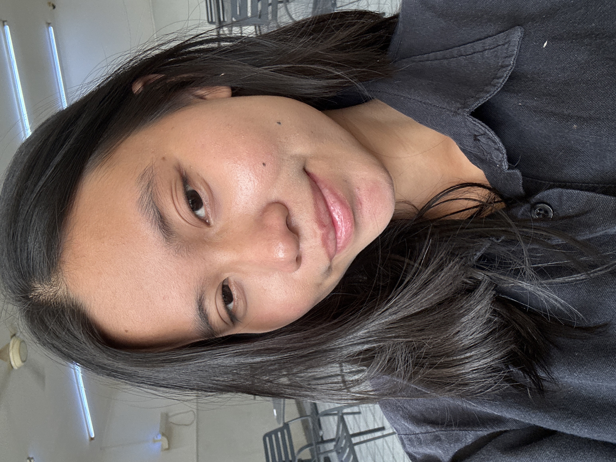 A young East Asian woman with long, dark hair is pictured in a close-up selfie. She is wearing a dark collared shirt. Her expression is neutral, with her lips slightly parted. The background suggests an indoor setting with fluorescent lighting and ceiling fans visible. There are also chairs and tables in the blurred background, indicating a common area or cafe. The image appears to have been taken in a public space within East Jakarta, Indonesia.