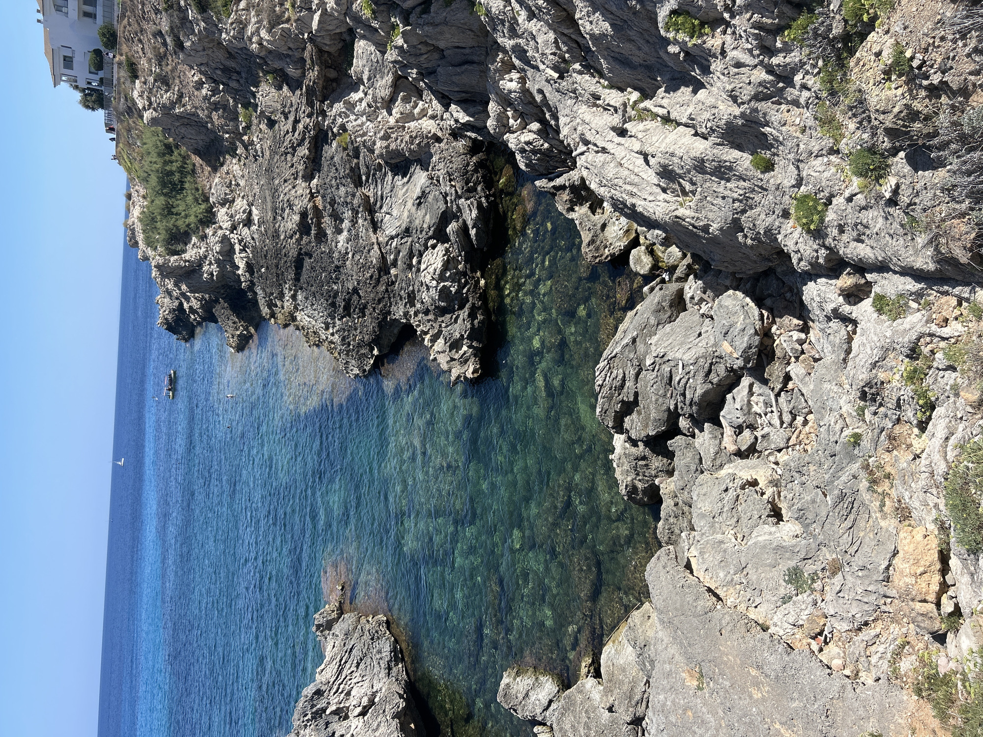 This image depicts a rocky coastline in Cala Lliteres, Spain. The foreground is dominated by a rugged, sun-drenched cliff face composed of light gray, weathered rocks and sparse vegetation. The rocks show intricate textures and formations, with some areas appearing darker, possibly indicating dampness or different mineral composition.

To the left, a small, clear turquoise cove is visible. The water is remarkably transparent, revealing the rocky seabed beneath, with some areas of lighter sand. The water is calm and shallow within the cove.

Beyond the cove, the sea stretches out, transitioning to a deeper blue further from the shore. A small white sailboat is visible on the horizon, and a yellow boat with people on board is closer to the coast, near the rocky shore.

On the upper right side of the image, a white building with a tiled roof is situated on a hill overlooking the sea. Several individuals, their details indistinct, are visible near the building, suggesting a residential or resort area. The sky is a clear, bright blue, indicating a sunny day. The overall impression is one of a scenic, Mediterranean coastal landscape.