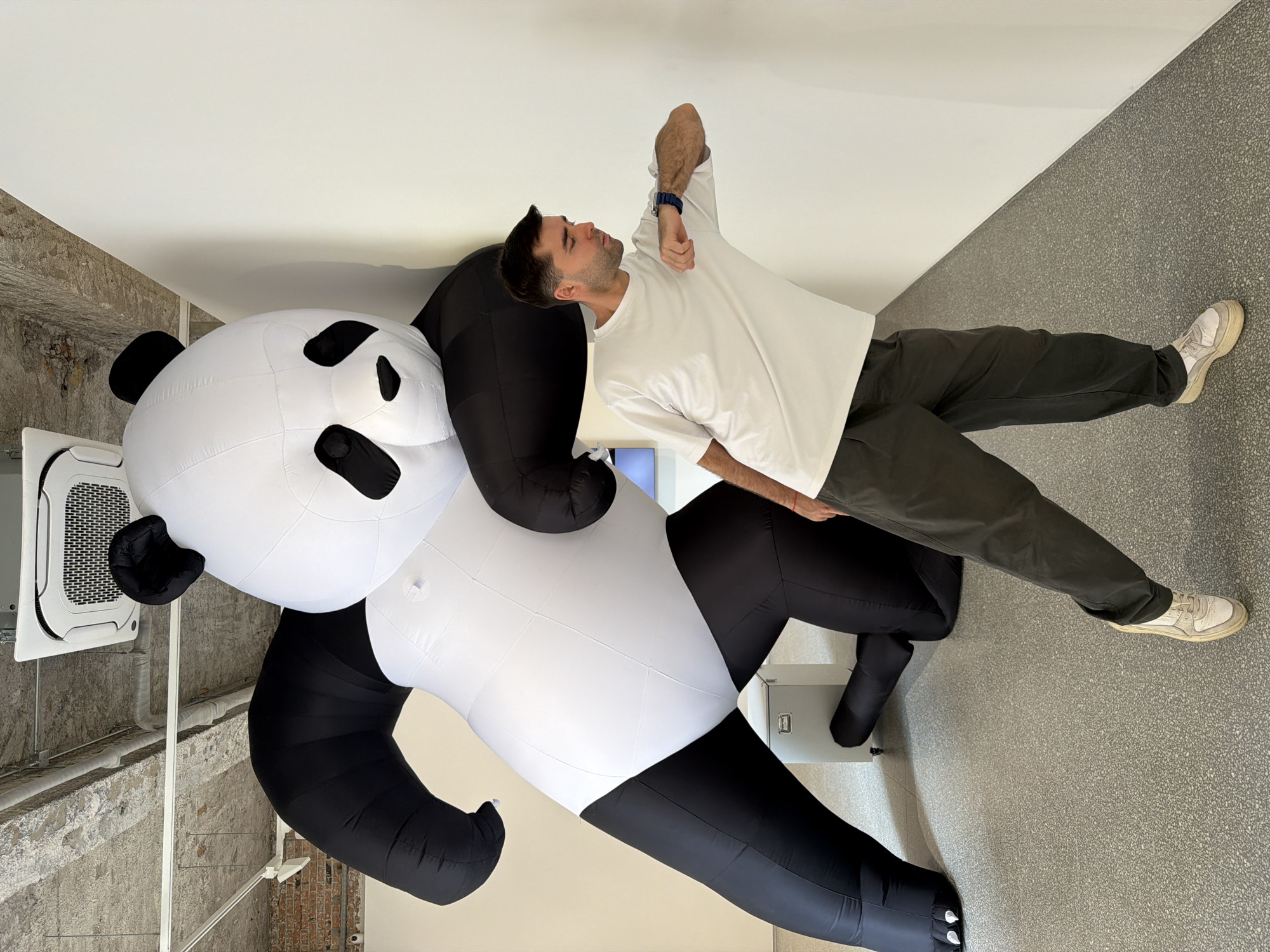 A man poses with an inflatable panda in an indoor space. The man is wearing a white t-shirt, gray pants, and white sneakers. He is standing with one arm raised. The inflatable panda is black and white, and it is larger than the man. The room has a gray speckled floor and a white ceiling. The location is in the city of Chaoyang District, China.