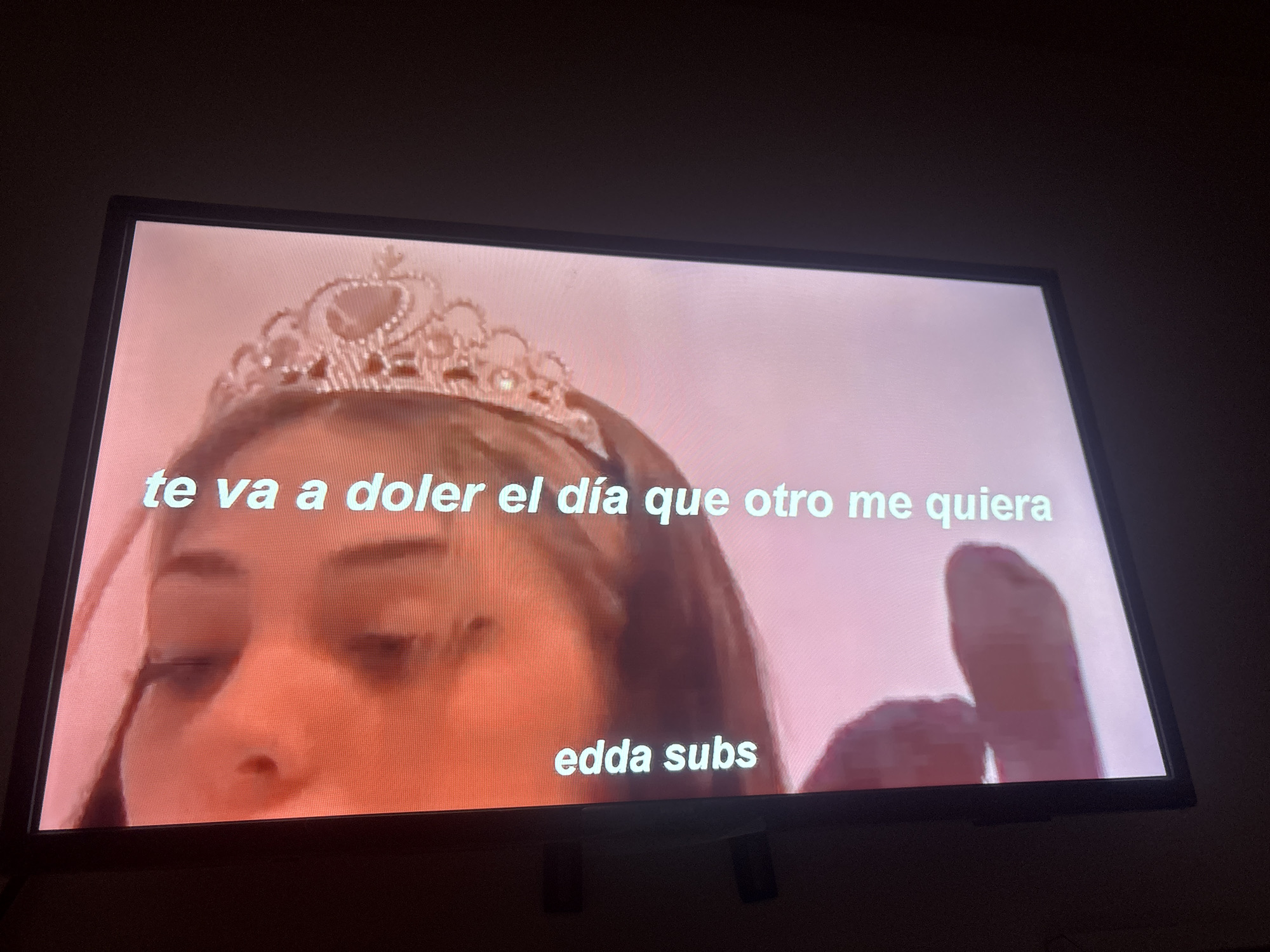 An image captures a photograph of a TV screen displaying a close-up of a young woman wearing a sparkling tiara on her head. The screen's background is a soft, light pink or peach color. The woman's face is visible from the nose up, with her eyes cast downwards, making her expression unclear. Her hair is dark, and her skin appears fair. To the right side of the screen, a large, indistinct, flesh-toned shape, possibly a hand or finger, is visible. Overlaid on the image are two lines of white text. The main text, "te va a doler el día que otro me quiera," translates from Spanish to "it will hurt you the day someone else loves me." Below this, smaller text reads "edda subs," likely indicating subtitle credits. The TV screen is encased in a dark frame, and the surrounding environment is dim, suggesting an indoor setting in low light, possibly evening or night. The Spanish text is consistent with the provided location context of Buenos Aires, Argentina.
