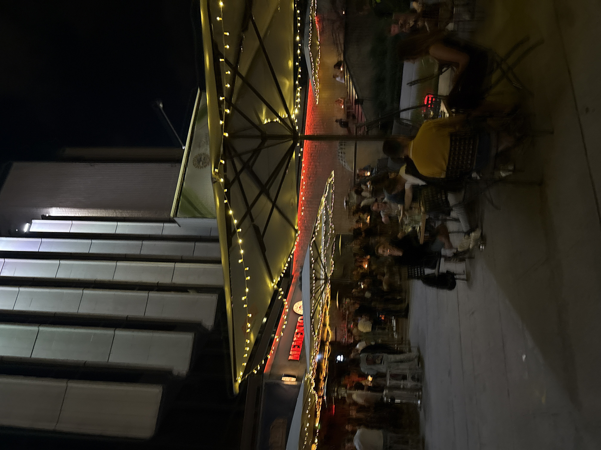 This image captures an outdoor evening scene at Memento NDK, a cafe and bar located at pl. "Bulgaria" 1 in Sofia, Bulgaria.
The scene depicts a bustling outdoor terrace illuminated by numerous string lights draped over large patio umbrellas. In the upper background, a tall, multi-story building with prominent vertical grey panels is visible, characteristic of the National Palace of Culture complex. The sky above is dark, indicating nighttime.
The central feature is the cafe's seating area, which is populated by numerous individuals. A bright red neon sign spelling "MEMENTO" is clearly visible above a section of the outdoor wall. Below this, the terrace features multiple levels, with steps leading to an elevated section where several people are seated.
People are distributed throughout the paved outdoor area, sitting at various tables and standing in groups. Objects observed include metal chairs, dark tables, tall silver outdoor heaters, and glasses on tables. One man in a yellow shirt is seated with his back to the viewer, facing another individual. To the right, another person is seated, smoking a cigarette, with a drink on their table. Many individuals are engaged in conversation or observing their surroundings. The overall ambiance is lively, consistent with a social gathering at a bar or cafe. The foreground consists of a dark, paved surface with subtle reflections.