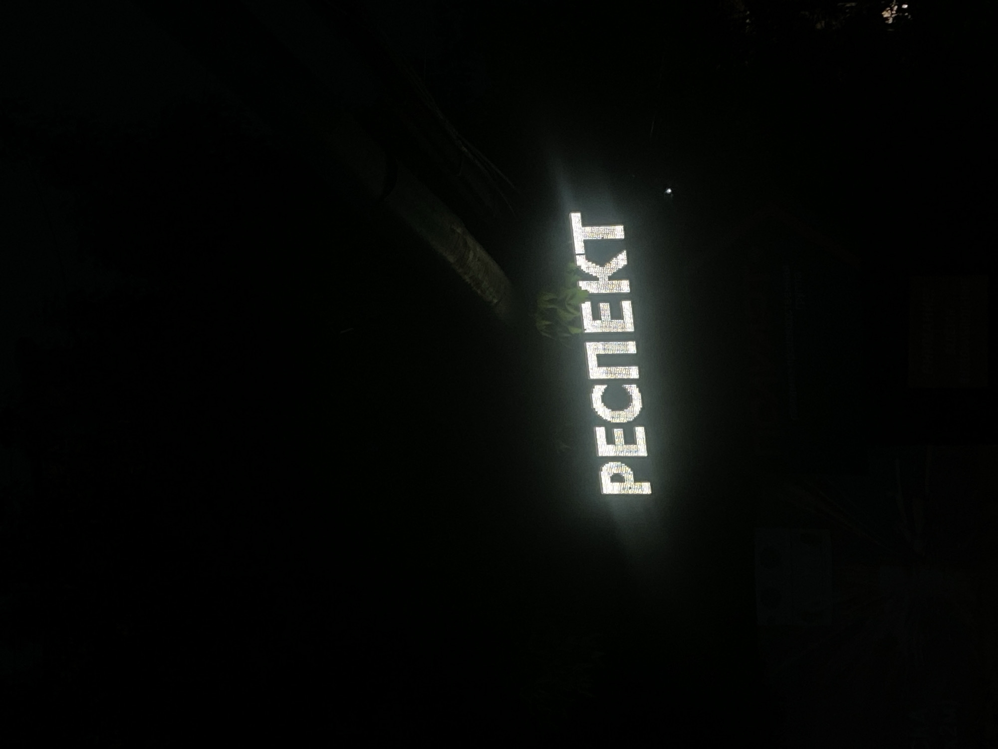 A nighttime shot shows a vertically oriented illuminated sign reading "РЕСПЕКТ" (RESPECT) in Cyrillic letters. The location is Rusanovo, Russia. The sign appears to be made of individual lights, creating a dotted effect. The background is dark, with some vague shapes suggesting trees or other structures.