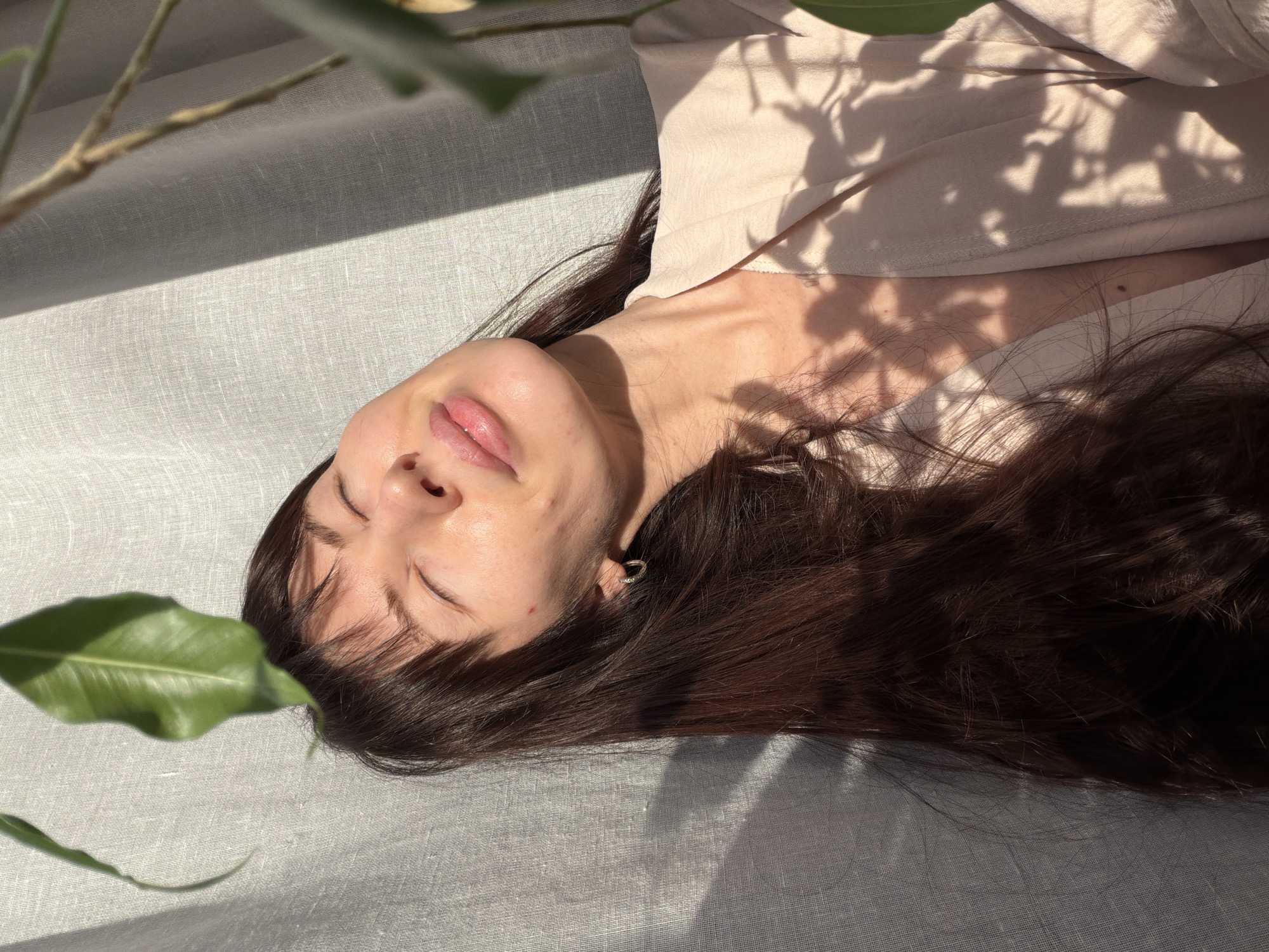 A woman with brown hair is lying down on a gray surface, possibly linen, in an indoor setting. She is wearing a light-colored shirt. Her eyes are closed and her face is illuminated by sunlight filtering through leaves, casting shadows on her face and clothing. A plant with large green leaves is visible in the lower left corner of the frame. The location, Perm, Russia is not discernible from the image.