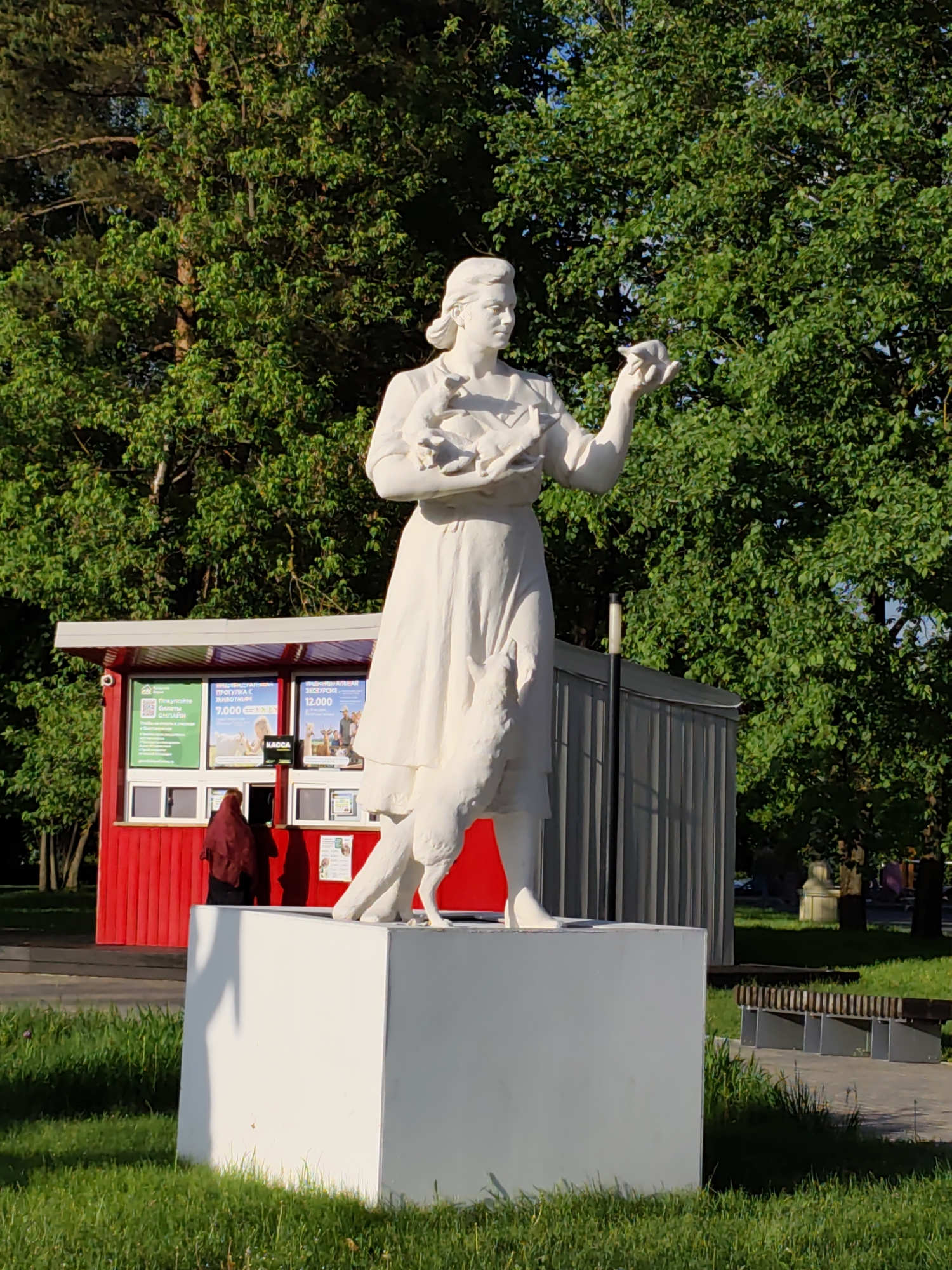 The image shows a white statue of a woman on a white pedestal. She is holding birds in her arms and one in her open hand. Behind her is a red and grey ticket kiosk where a person in a red scarf is buying a ticket. Green trees fill the background. A bench and pavement can be seen to the right of the statue. The location is the Moscow Zoo in Moscow, Russia.