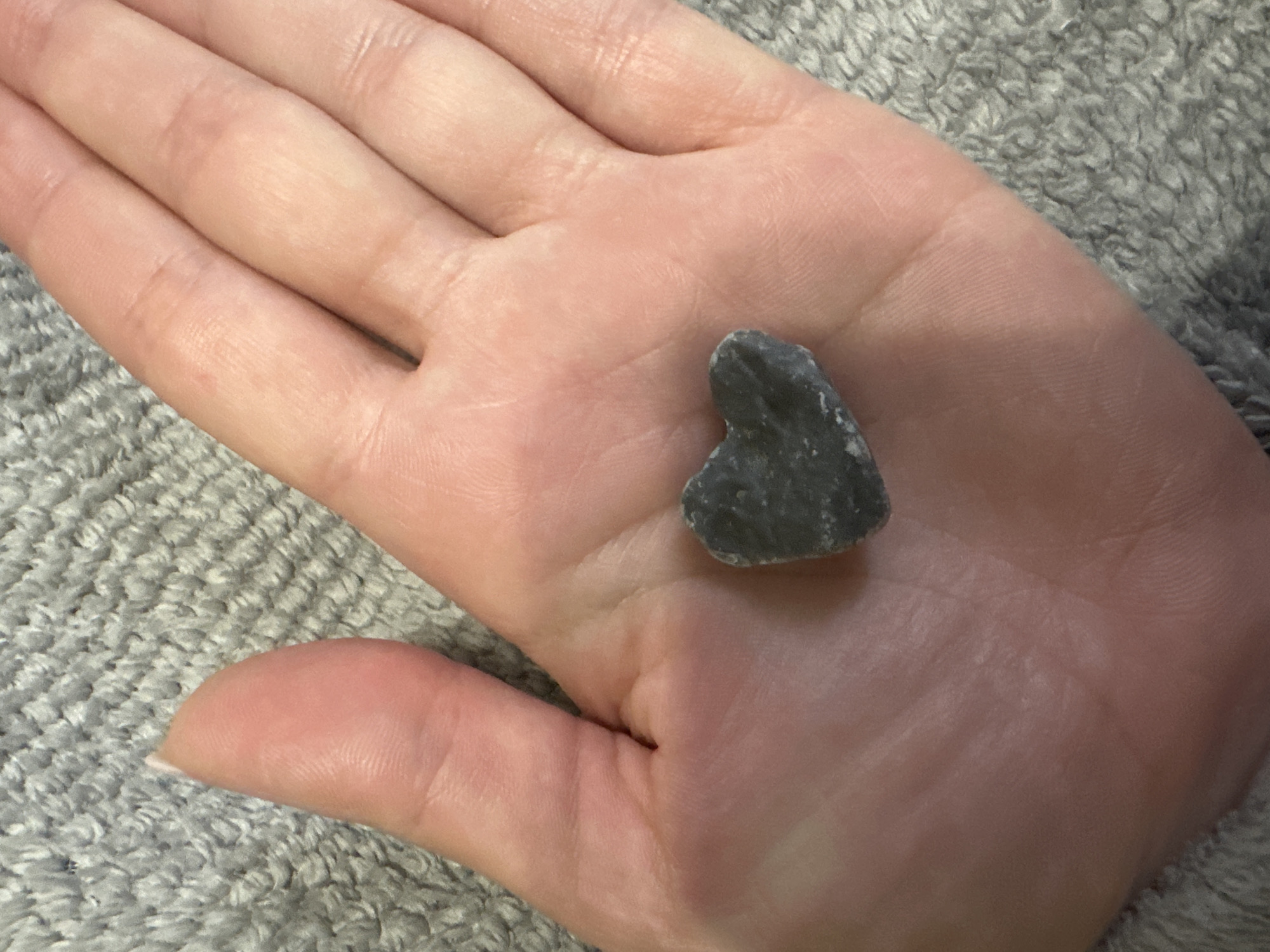 A close-up, top-down photograph captures a person's left hand presenting a small, dark grey, heart-shaped stone. The hand, with a fair skin tone, exhibits clear palm lines and finger creases. The visible thumb on the left side of the frame features a clean, light-colored fingernail. The stone, centrally placed in the palm, is irregular in shape, suggestive of a natural pebble, and displays variations in its dark grey hue with lighter grey specks and an uneven surface. The background is a uniformly textured grey fabric, likely a towel or blanket, characterized by a soft, looped pile. The scene is lit by soft, even illumination. The location is in the city of Unknown, Unknown.