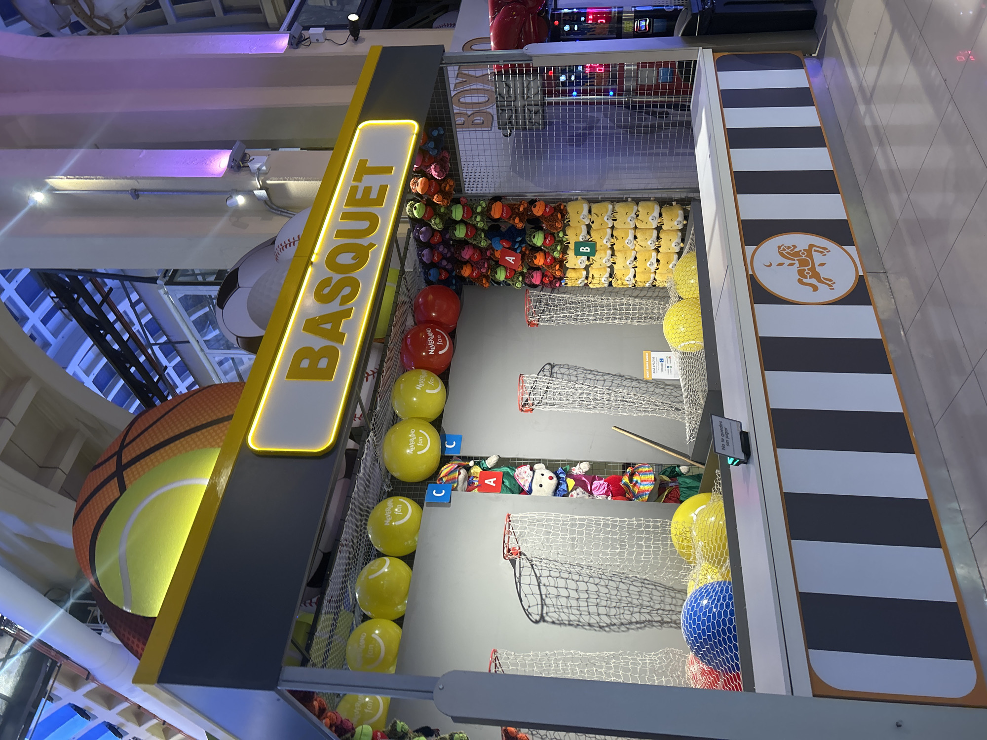 This image captures a "Basquet" (basketball) themed carnival game stall, likely located in a mall or entertainment complex in Buenos Aires, Argentina, given the signage and general ambiance. The stall itself is brightly lit, with a yellow illuminated sign displaying the game's name. Above the stall, a large decorative structure resembling a combination of a tennis ball and a basketball is visible.
The game involves throwing balls into netted hoops. Several yellow balls with the "Neverland Fun" logo are displayed along the top edge of the stall, some appearing to be inside mesh bags. Below these, a collection of plush toys, including stuffed monkeys and possibly other animals, are hung as prizes, organized into sections labeled "A" and "B." The lower section of the stall contains several larger balls, including one blue, one red, and two yellow balls, also within mesh.
To the right of the basketball game, another stall is partially visible, featuring large red boxing gloves and a sign that appears to say "BOXO." The overall environment suggests an indoor, brightly lit recreational area. The flooring is tiled, and the background shows architectural elements characteristic of a modern building, including columns and ceiling structures with lighting. No people are actively engaged in playing the game in this particular frame.