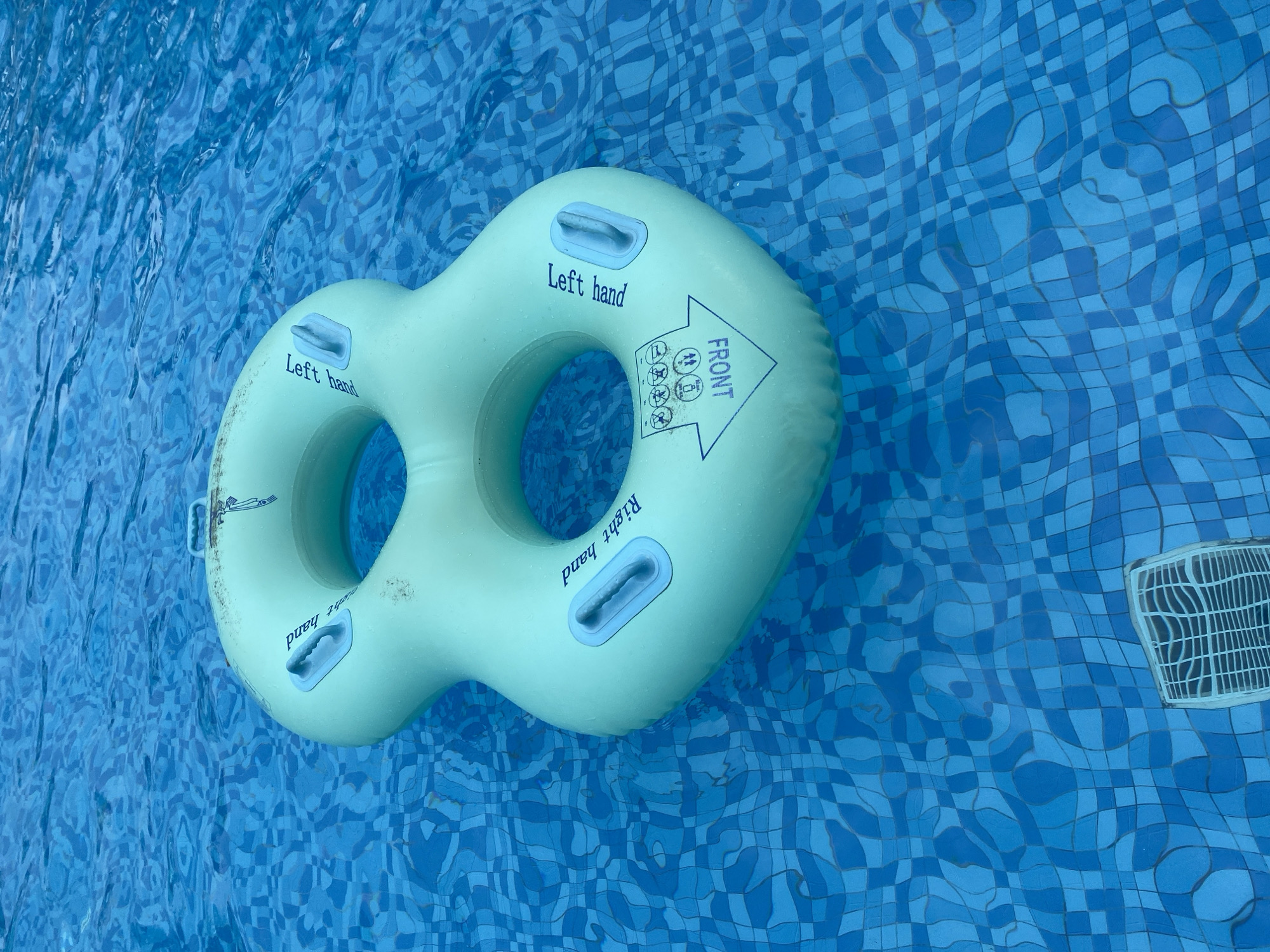 A light green, figure-eight shaped inflatable pool float is floating in a swimming pool with blue tile. The float is oriented diagonally in the frame. Text on the float reads "Left hand", "Right hand", and "FRONT" along with icons representing prohibited activities. A pool drain is visible in the bottom right corner of the frame. The scene is located in Cam Lam, Vietnam.