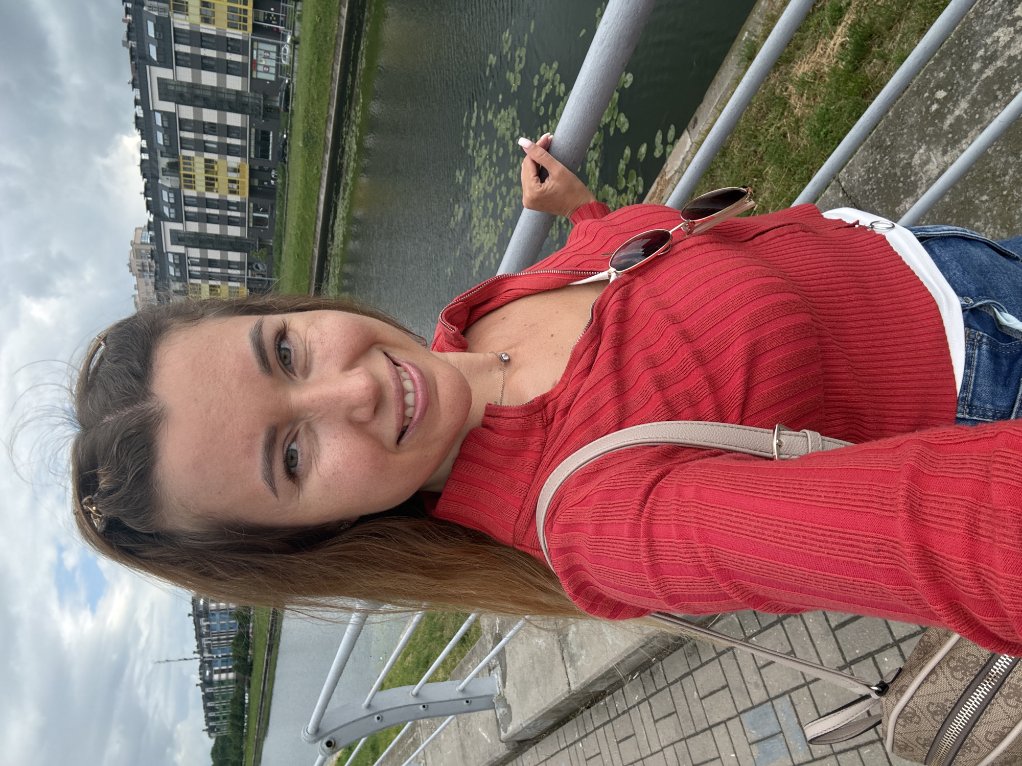 A woman takes a selfie outdoors on a cloudy day, standing on a paved walkway next to a metal railing. The woman has long, brown hair pulled back in a ponytail and is wearing a red, ribbed zip-up top over a white garment, and blue jeans. She is smiling at the camera, and sunglasses are hanging from the neckline of her top. A patterned shoulder bag is visible on her left side. Behind her, a body of water with lily pads is bordered by a grassy embankment and a row of modern, multi-story buildings.