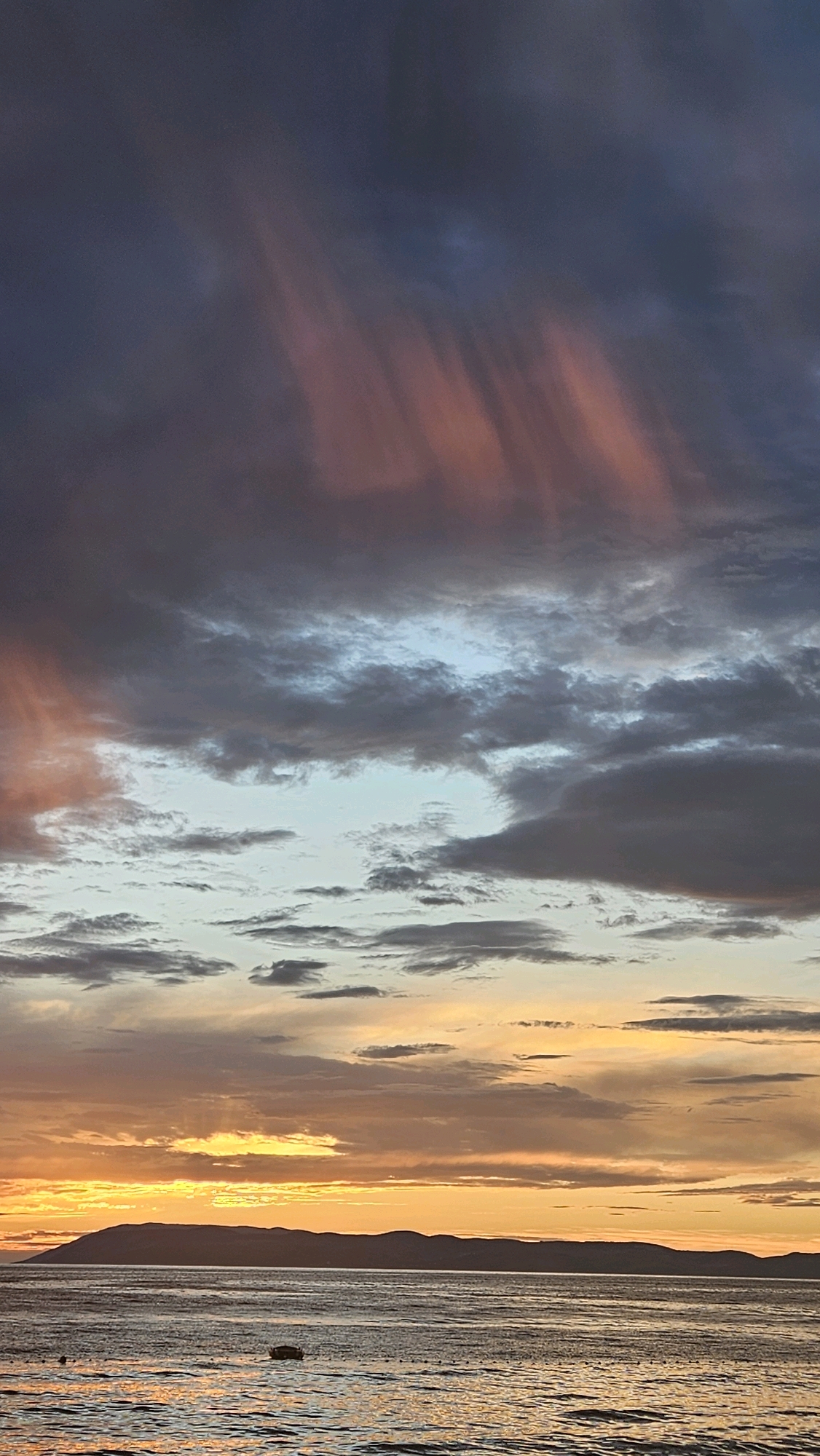 This is a vertical, eye-level, medium shot photograph of a sunset over the Adriatic Sea, likely taken from the coast of Općina Tučepi, Croatia. The sky is filled with a dramatic display of clouds, with hues of deep gray, purple, and streaks of pink and orange. The sun is low on the horizon, casting a warm, golden glow across the water. In the distance, a dark, undulating silhouette of a mountain range is visible. The sea is choppy, reflecting the colors of the sky. A small, dark object, possibly a boat, is present on the water in the lower portion of the image. The overall mood is serene and visually striking, capturing the beauty of a coastal sunset.
