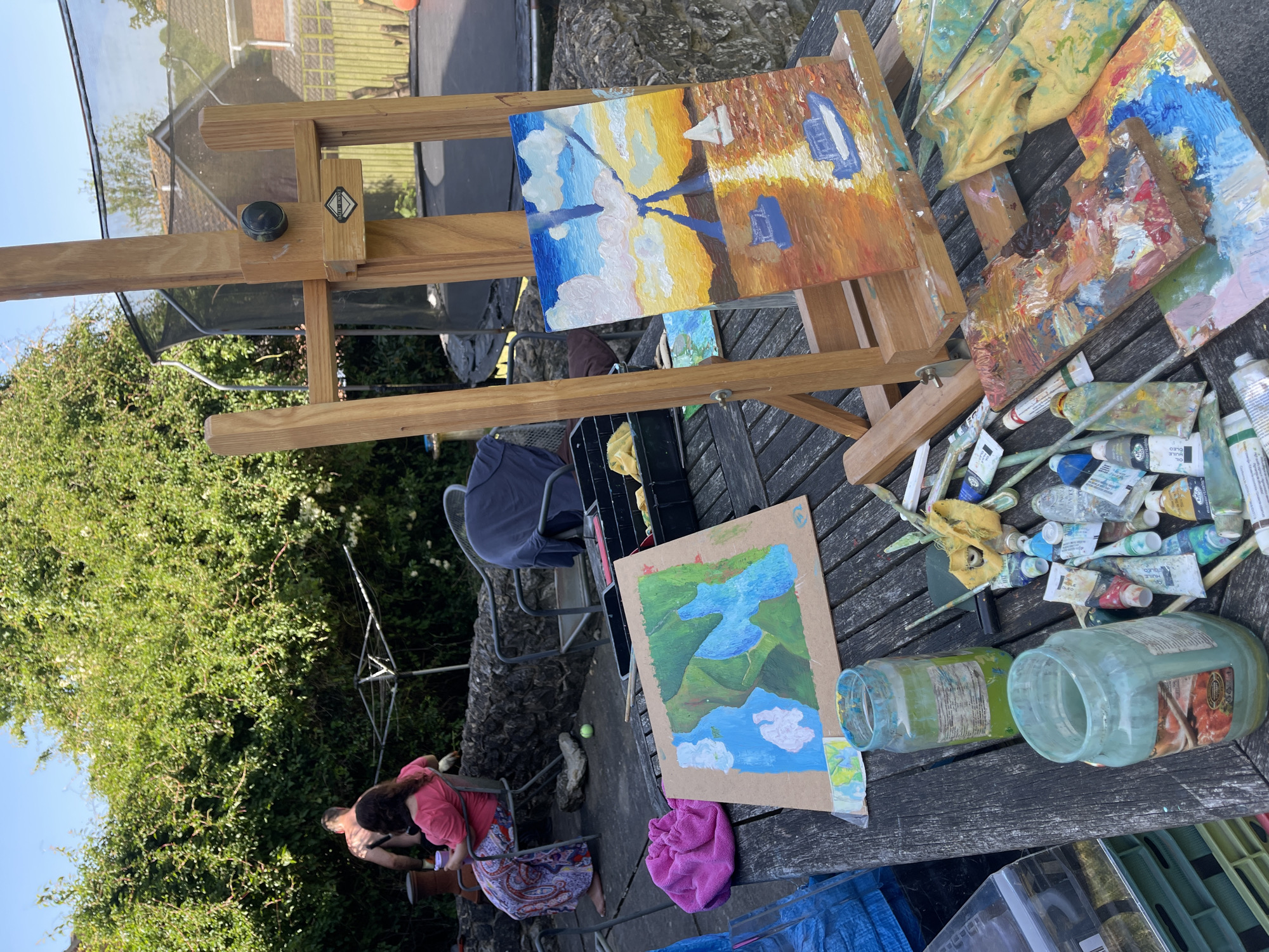 The image presents an outdoor art studio setting in Maidstone, United Kingdom. A wooden easel supports a partially completed painting depicting a coastal scene with boats and a colorful sky. Another painting lies flat on a wooden table, showing a landscape with water and mountains. The table is cluttered with paint tubes, brushes, a used palette covered in mixed paints, and glass jars, likely for cleaning brushes. In the background, partially obscured by foliage, two individuals are seated. One is wearing a pink shirt and patterned skirt. A clothesline is visible amidst the trees. The scene appears to be set in a garden or patio area.