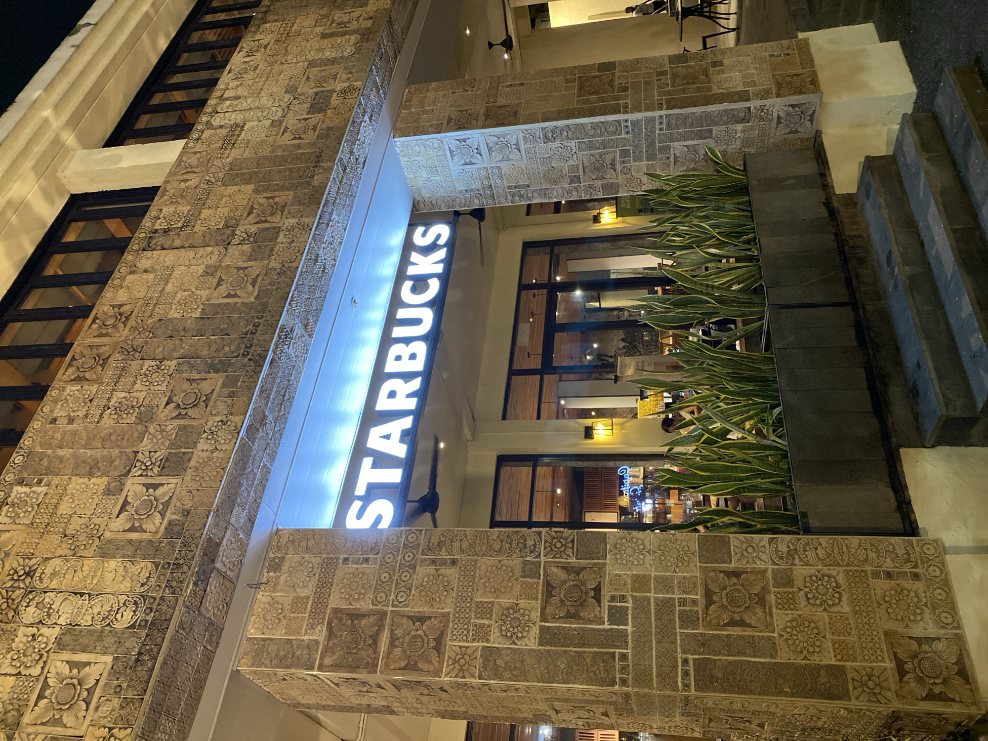 The image shows the exterior of a Starbucks in Kuta Selatan, Indonesia, at night. The building features ornate, carved stone tiles on its facade and pillars. A brightly lit "STARBUCKS" sign is prominently displayed above the entrance. In front of the store, there is a planter box filled with snake plants. The entrance has a set of stairs leading up to it, and a few tables and chairs are visible outside the establishment. The overall ambiance is illuminated by the store's internal lighting and decorative exterior lamps.