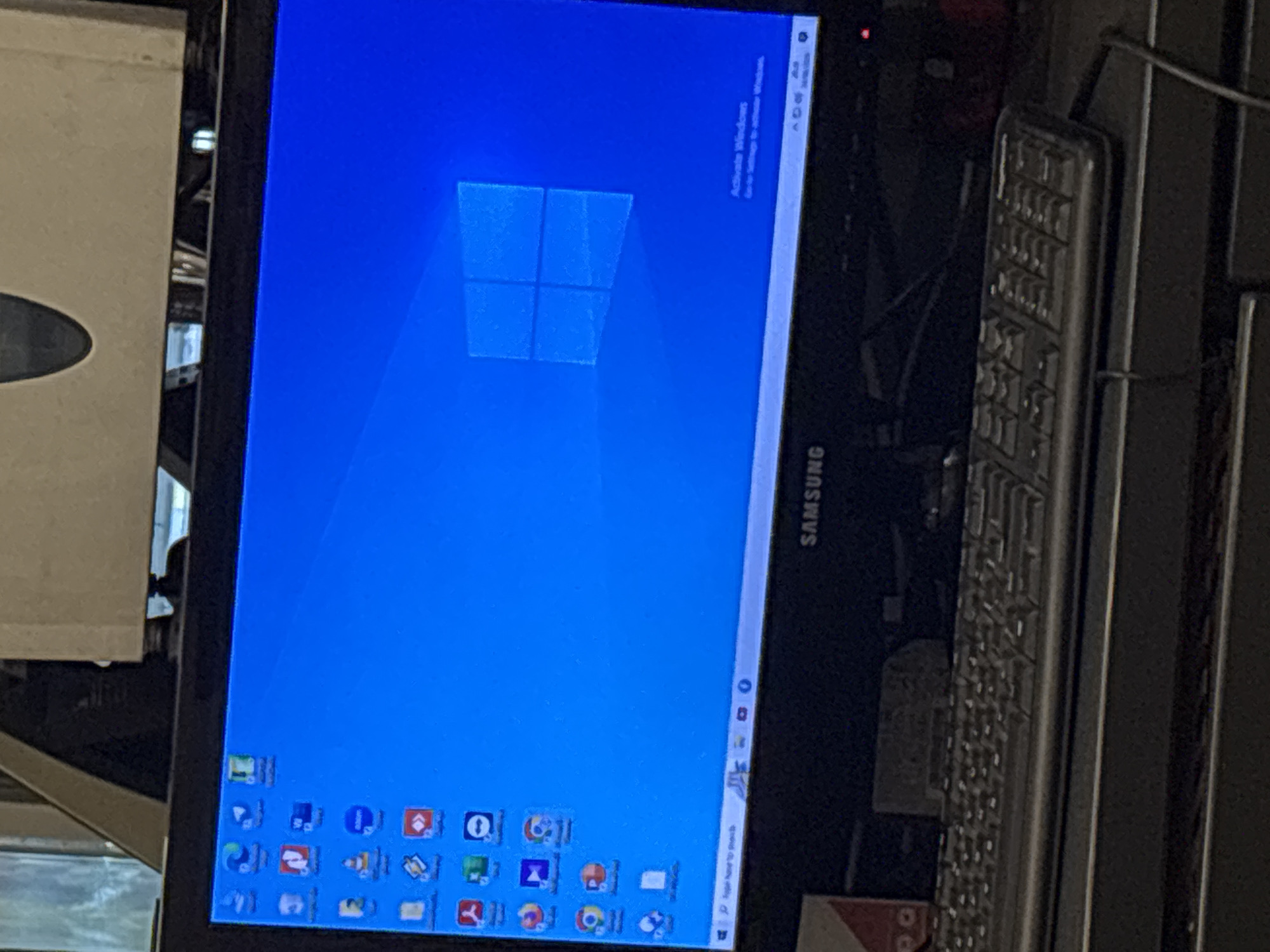 This is a close-up, eye-level shot of a computer monitor displaying the Windows 10 operating system. The screen is predominantly blue, featuring the default Windows logo wallpaper. On the left side of the screen, numerous application icons are arranged in columns. The bottom of the screen shows the Windows taskbar, with icons for open applications and system notifications in the lower right corner. Below the monitor, a black keyboard is visible, with some of its keys illuminated by the screen's glow. The setting appears to be an indoor workspace.
