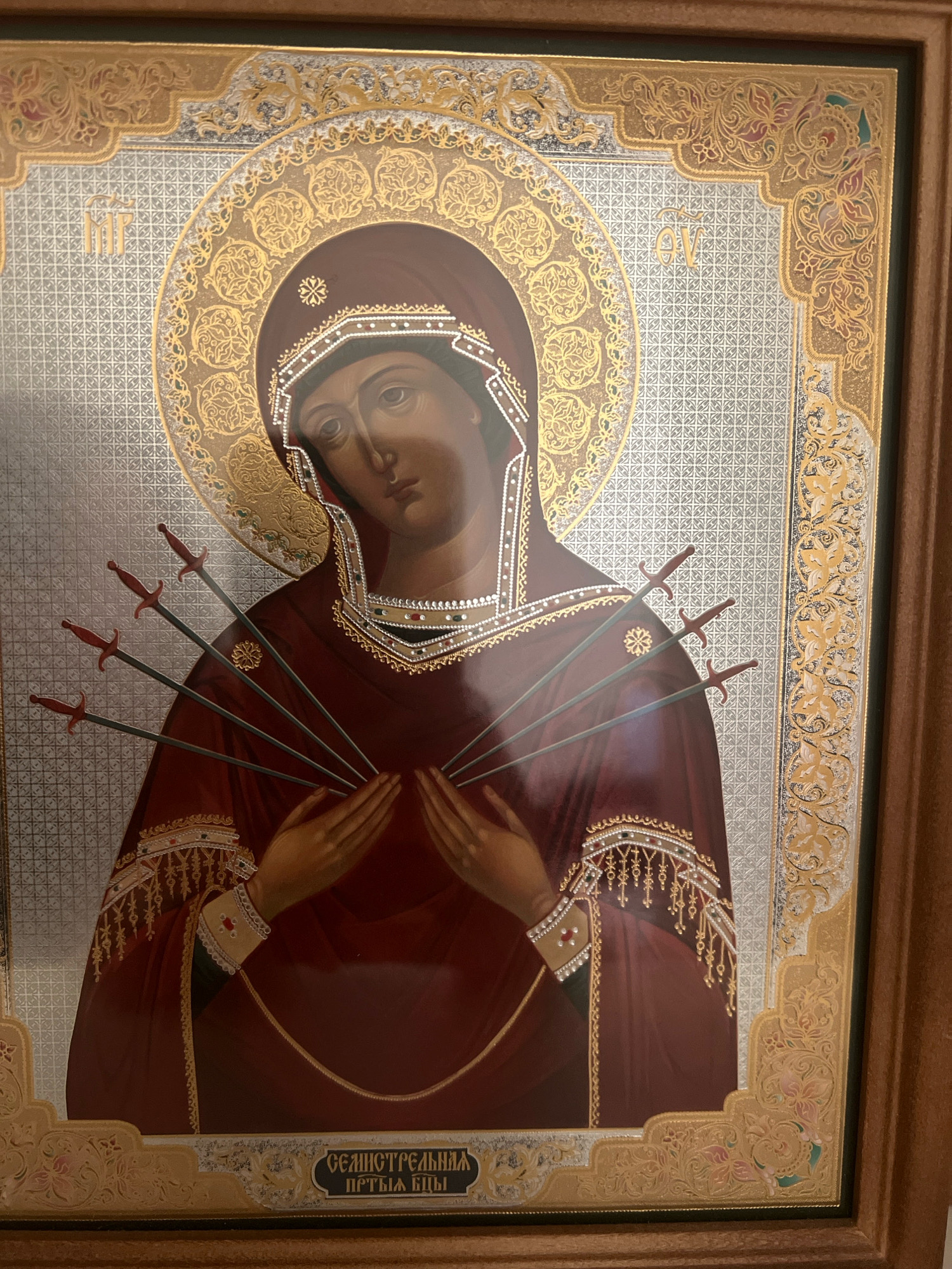 The image shows an icon of the Virgin Mary, depicted with seven swords piercing her heart, a symbol of her sorrow. The icon is framed in gold and features intricate decorative borders. In the lower section of the icon, there is text in Cyrillic script, which translates to "The Softener of Evil Hearts." The icon is placed on a wooden surface, likely a wall or a piece of furniture, and the overall setting appears to be indoors. The lighting is warm and diffused, suggesting it may be evening or that the room is dimly lit. There is no specific information within the image to determine its location in the city of Volzhsky, Russia.