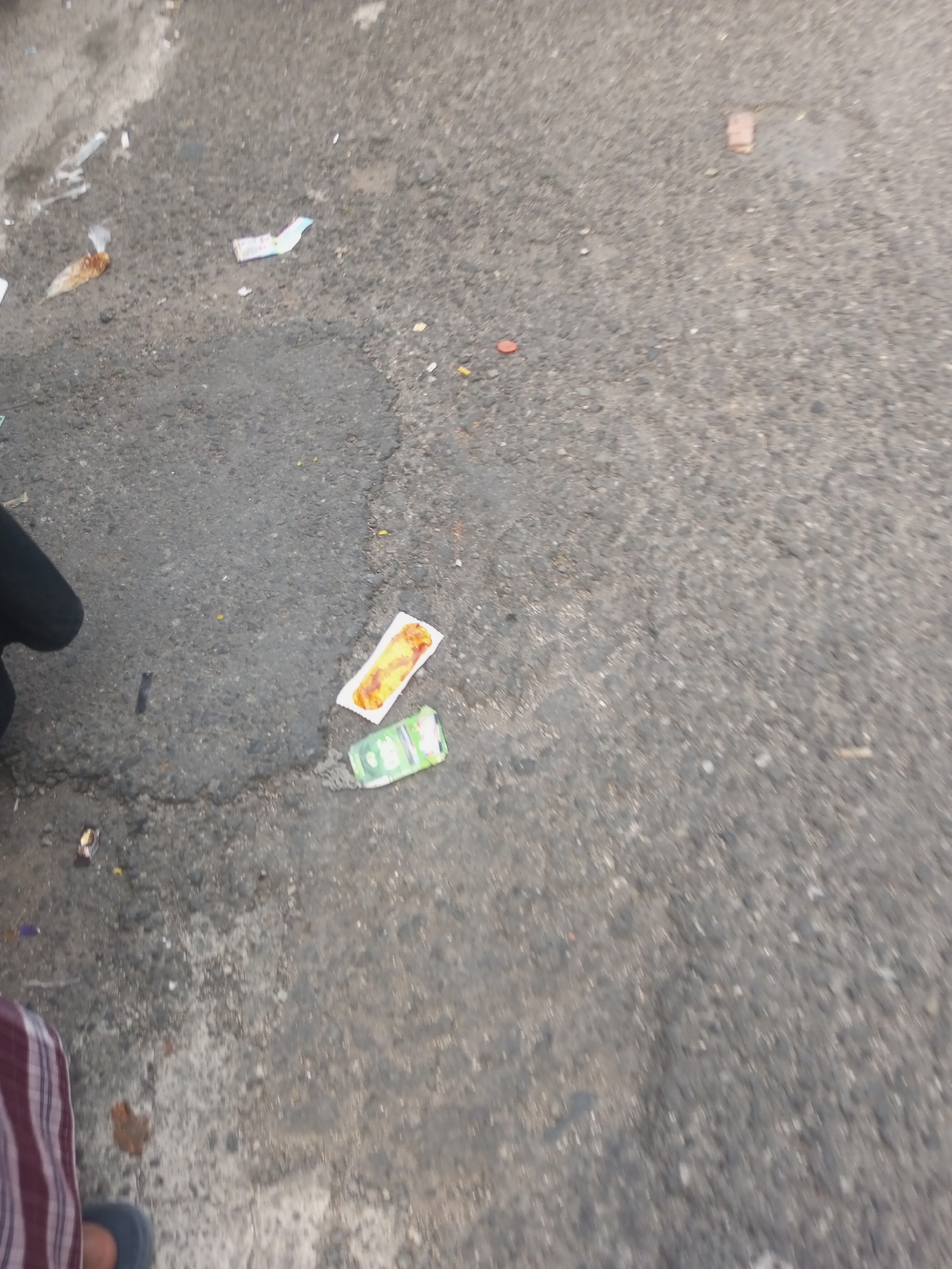 The image shows a close-up, slightly blurred view of a paved street in Pandansari, Indonesia. The ground is asphalt, with some patched areas and debris scattered across it. In the foreground, a person's leg, clad in striped fabric and a dark shoe, is visible on the left side of the frame. There are several pieces of litter on the ground: a crumpled wrapper, a small piece of paper with a food stain, and a green packet. Further back, other small pieces of debris and a single red object are visible. The lighting suggests it is daytime.